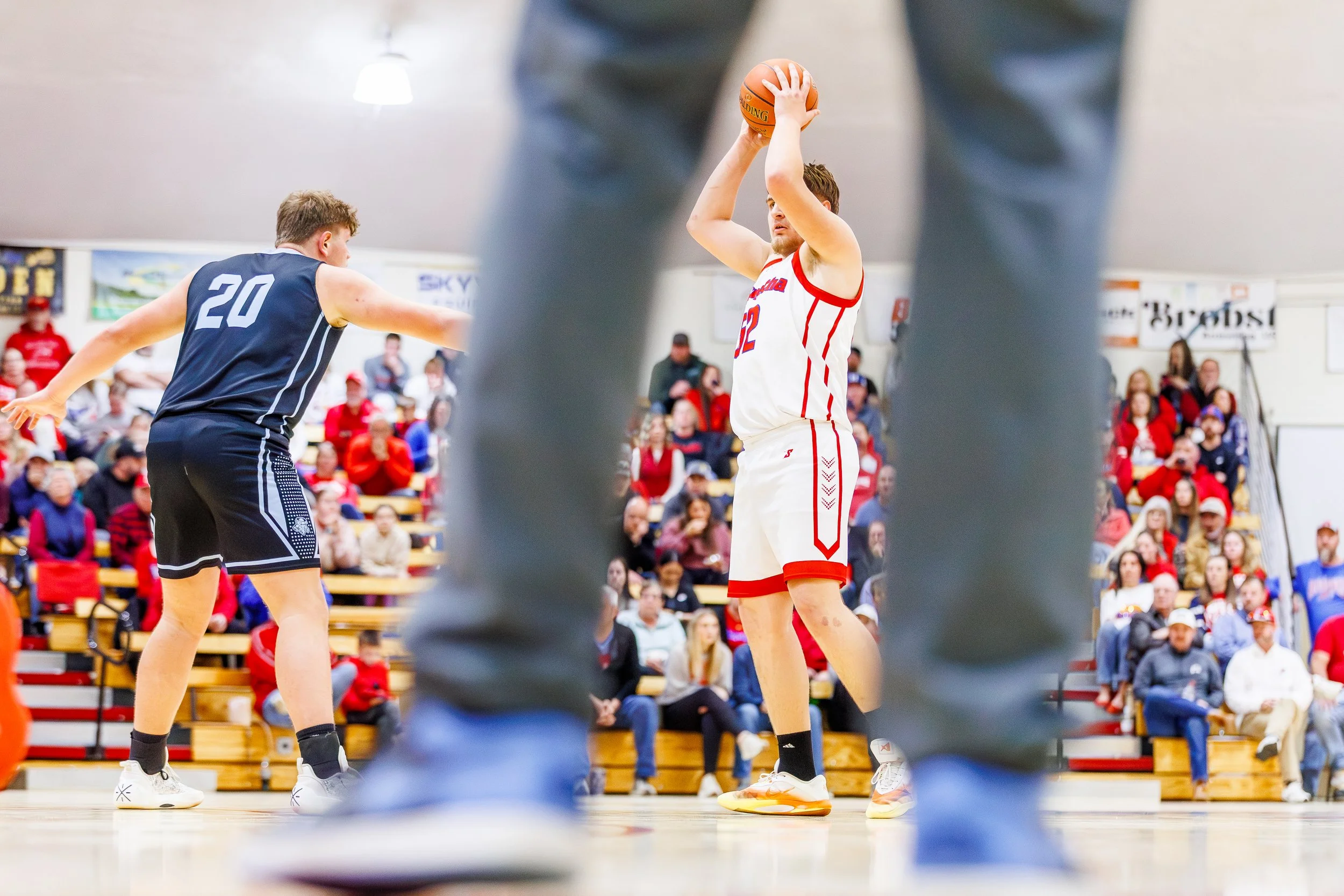 A basketball player in a white and red uniform preparing to shoot, seen through the legs of another player or spectator in the foreground, with an audience watching in the background.