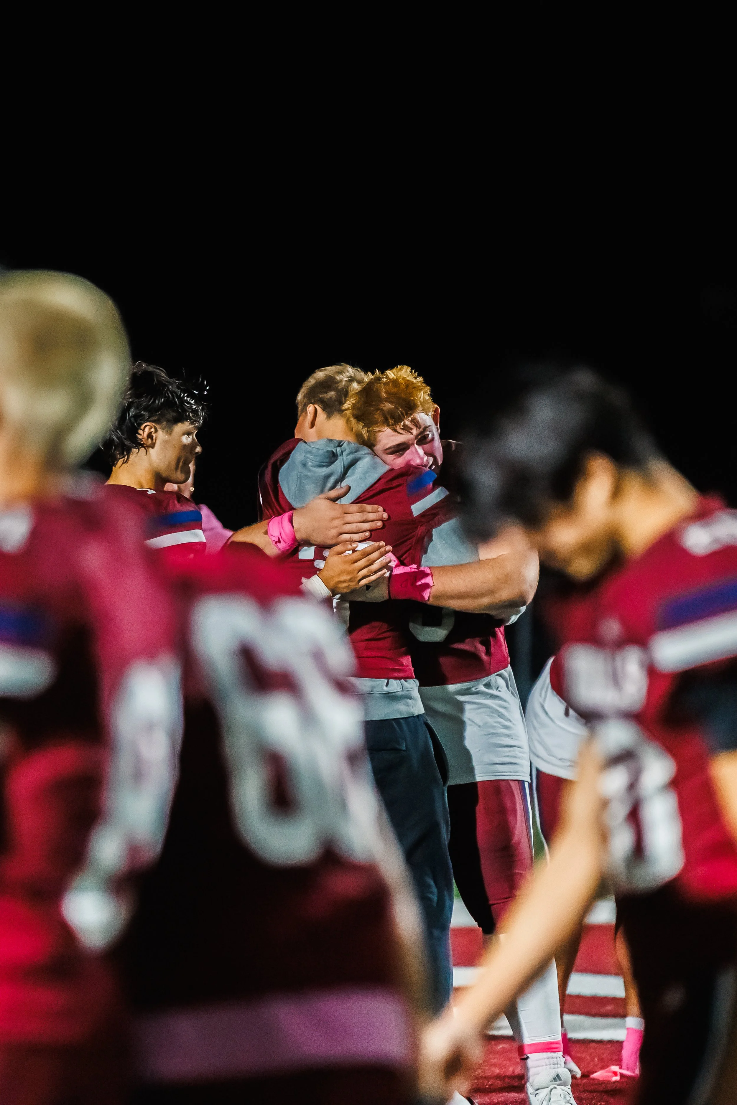 Football players hugging during a game at night, emotional celebration, wearing red and gray uniforms.