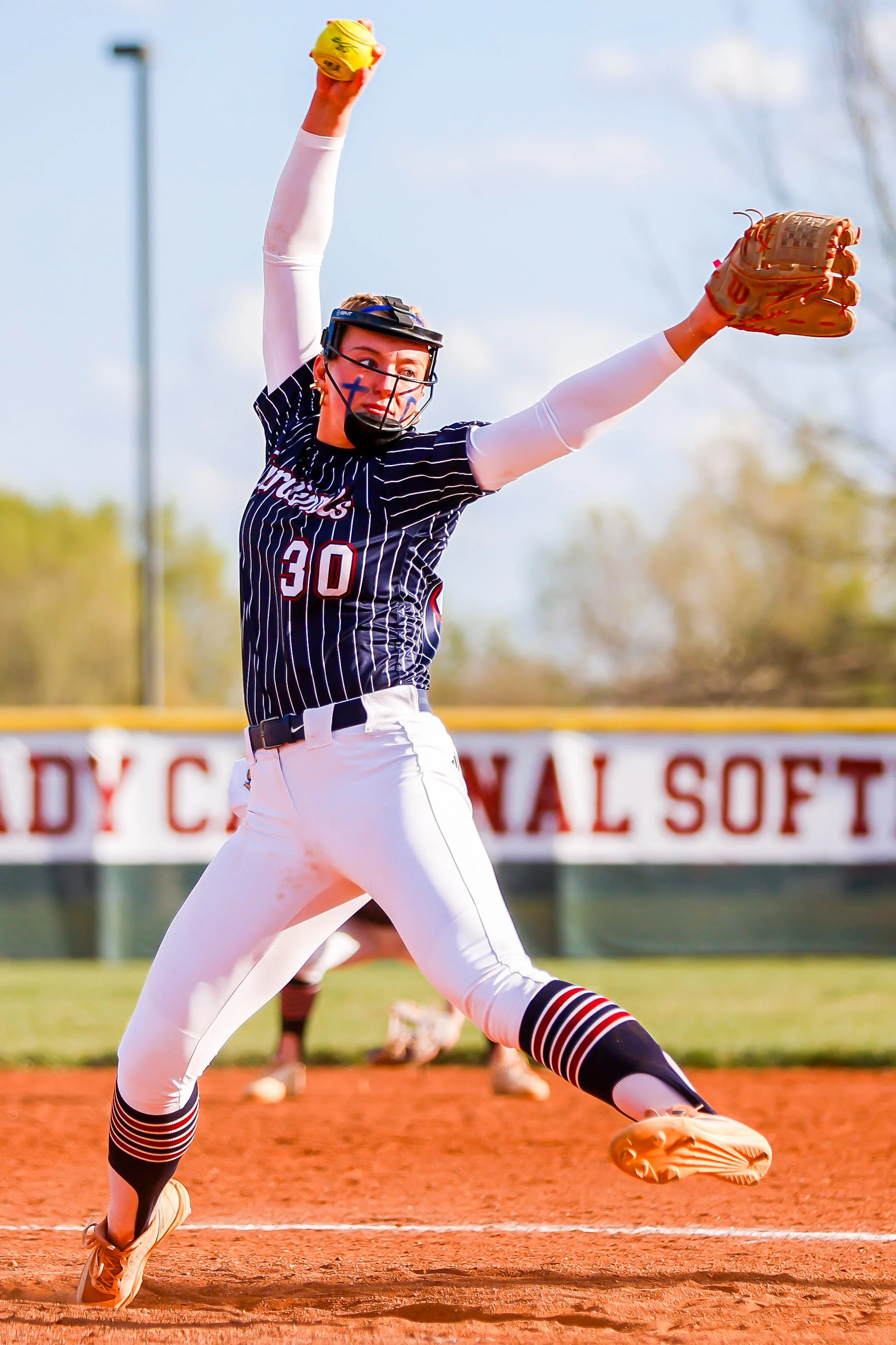 A female softball player in a black and white striped uniform with the number 30 is caught mid-celebration, raising her yellow glove and ball in the air on a softball field.