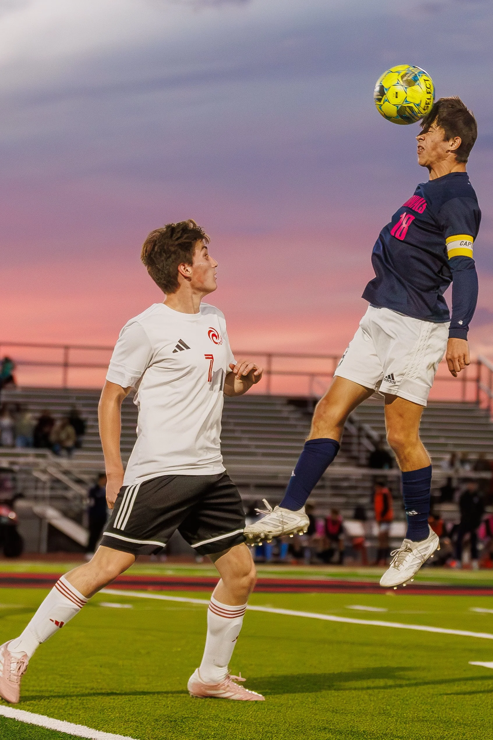 Two soccer players in action during a match at sunset, one jumping to head the ball while the other watches.