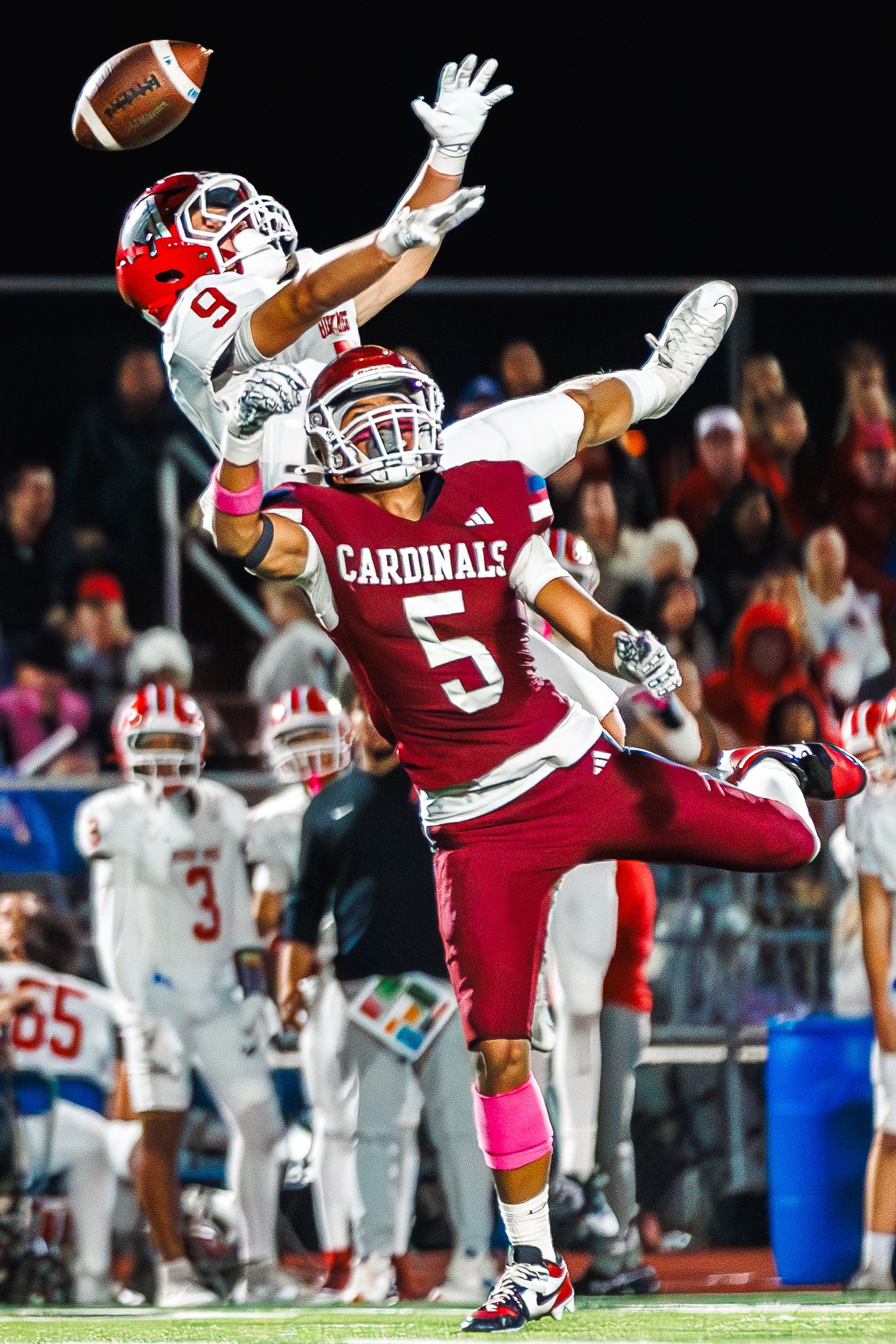An American football game showing a player in a red uniform with number 5, jumping with one leg raised, blocking an opposing player in a white uniform with number 9, who is trying to catch a football in mid-air. The background has spectators and othe
