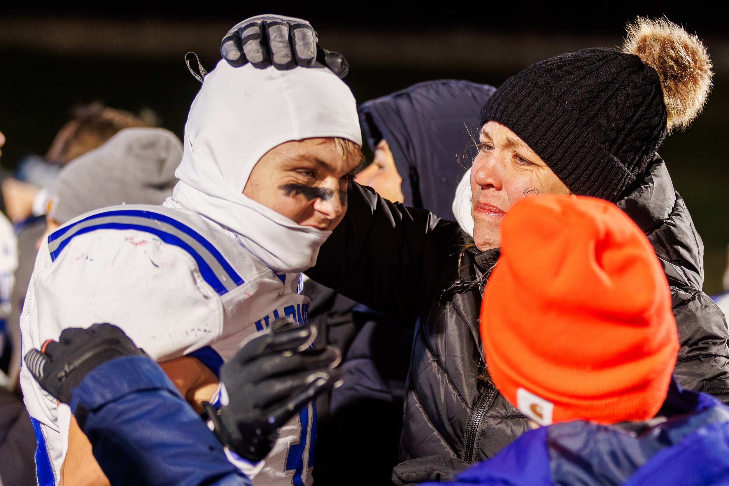 A football player in uniform and face paint is comforted by a woman in a black jacket and hat, while another person in an orange hat looks on.