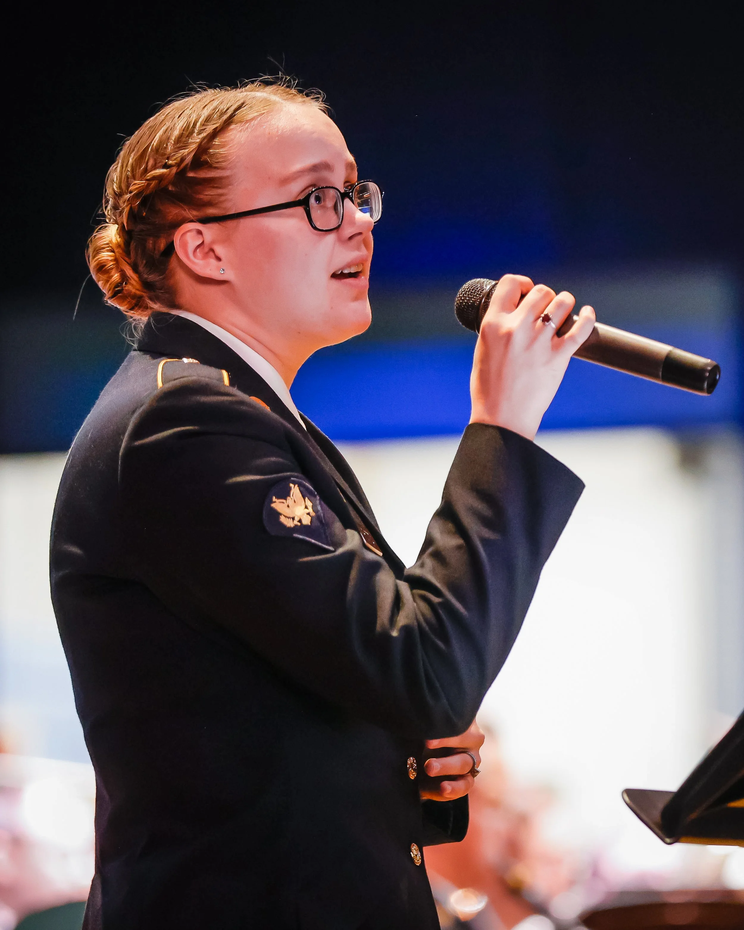 A woman in a military uniform with glasses is speaking into a handheld microphone at an event.