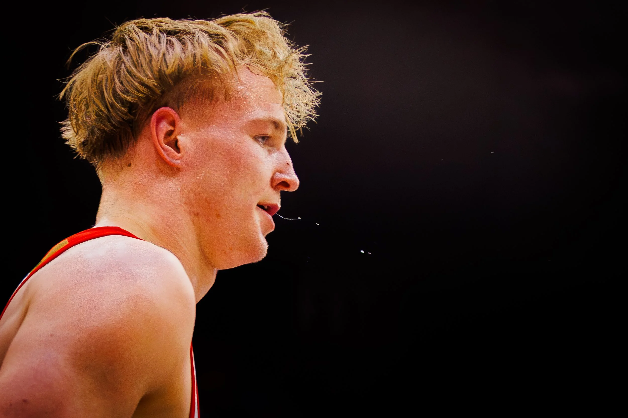 Close-up profile of a young male athlete with blond hair, sweating, against a black background.