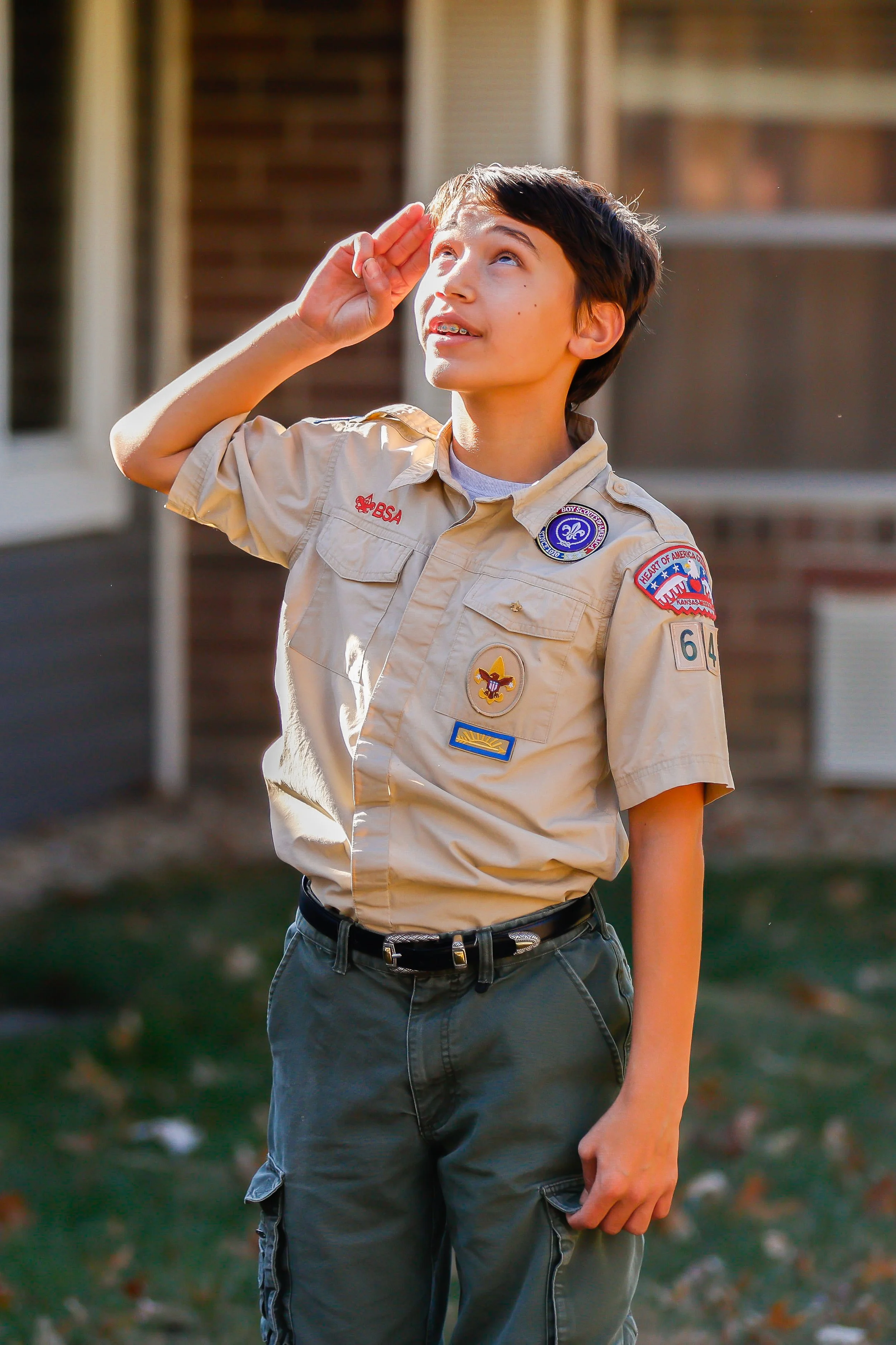 A Boy Scout saluting outdoors during daytime, wearing a tan Scout uniform with patches and badges, standing in front of a house with a porch and window.