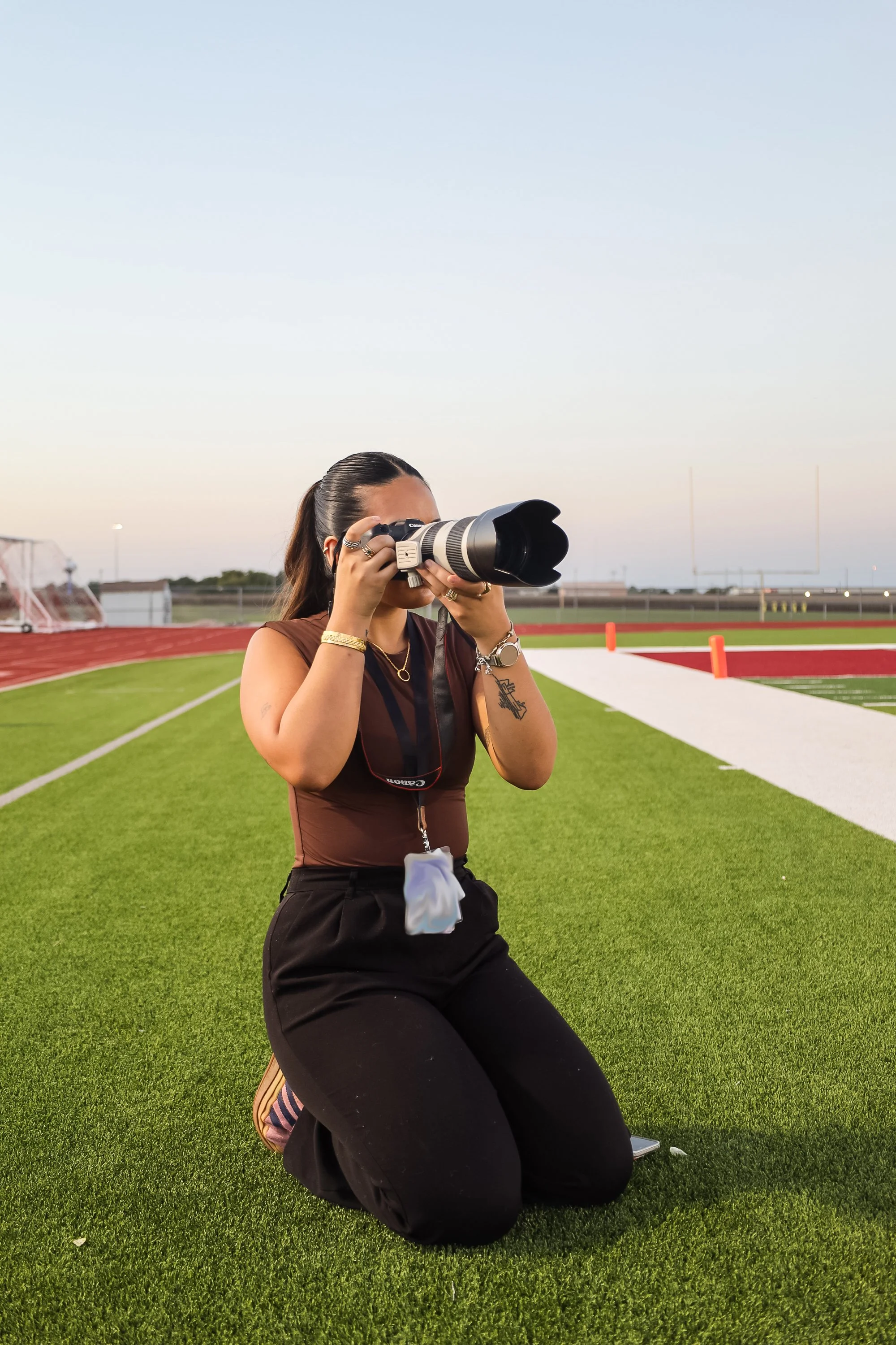 Woman kneeling on grass field, holding a professional camera with a large lens, taking photos on a sports field at dusk.