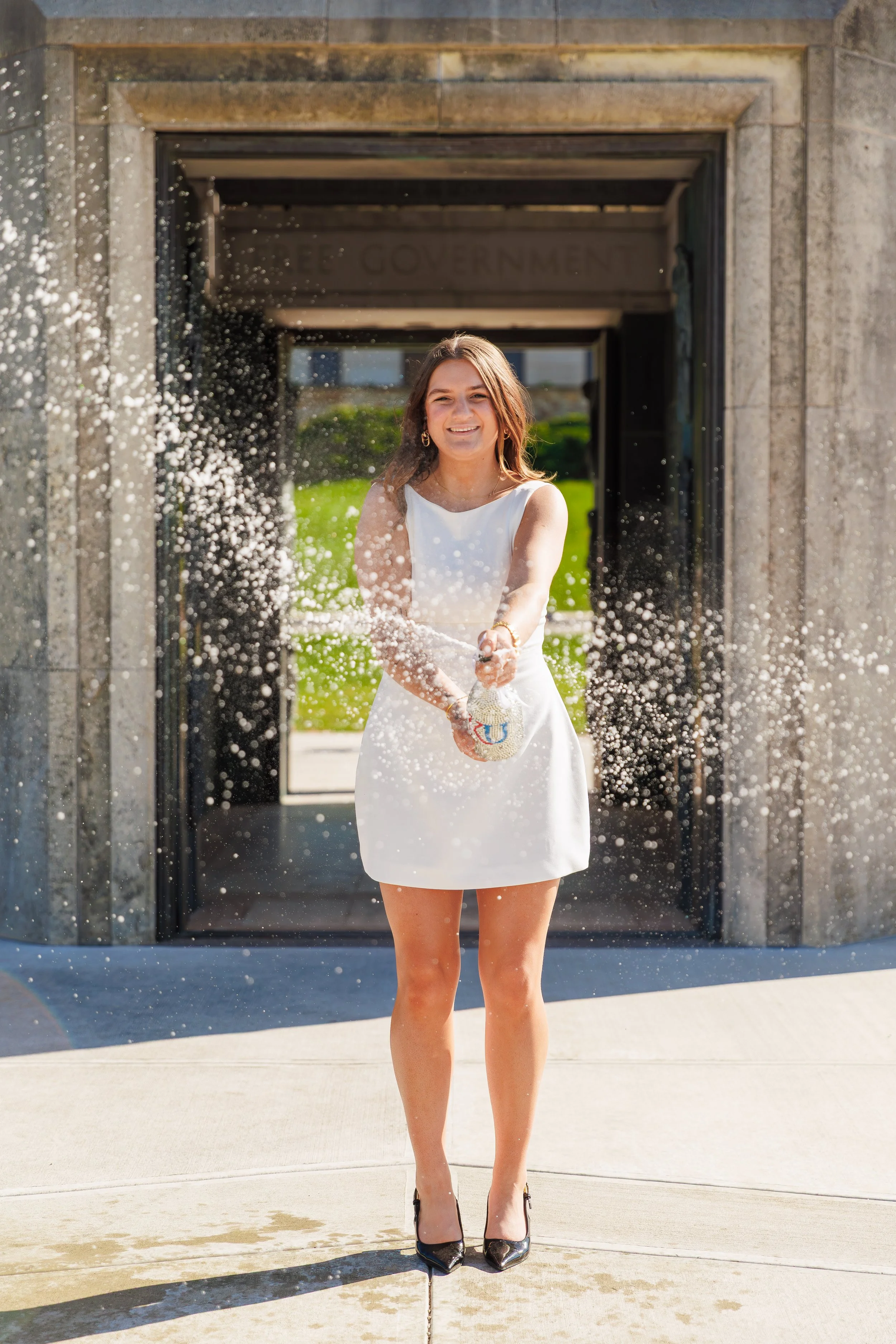 A young woman in a white dress is smiling and holding a purse as she stands in front of a government building, with water splashing around her.
