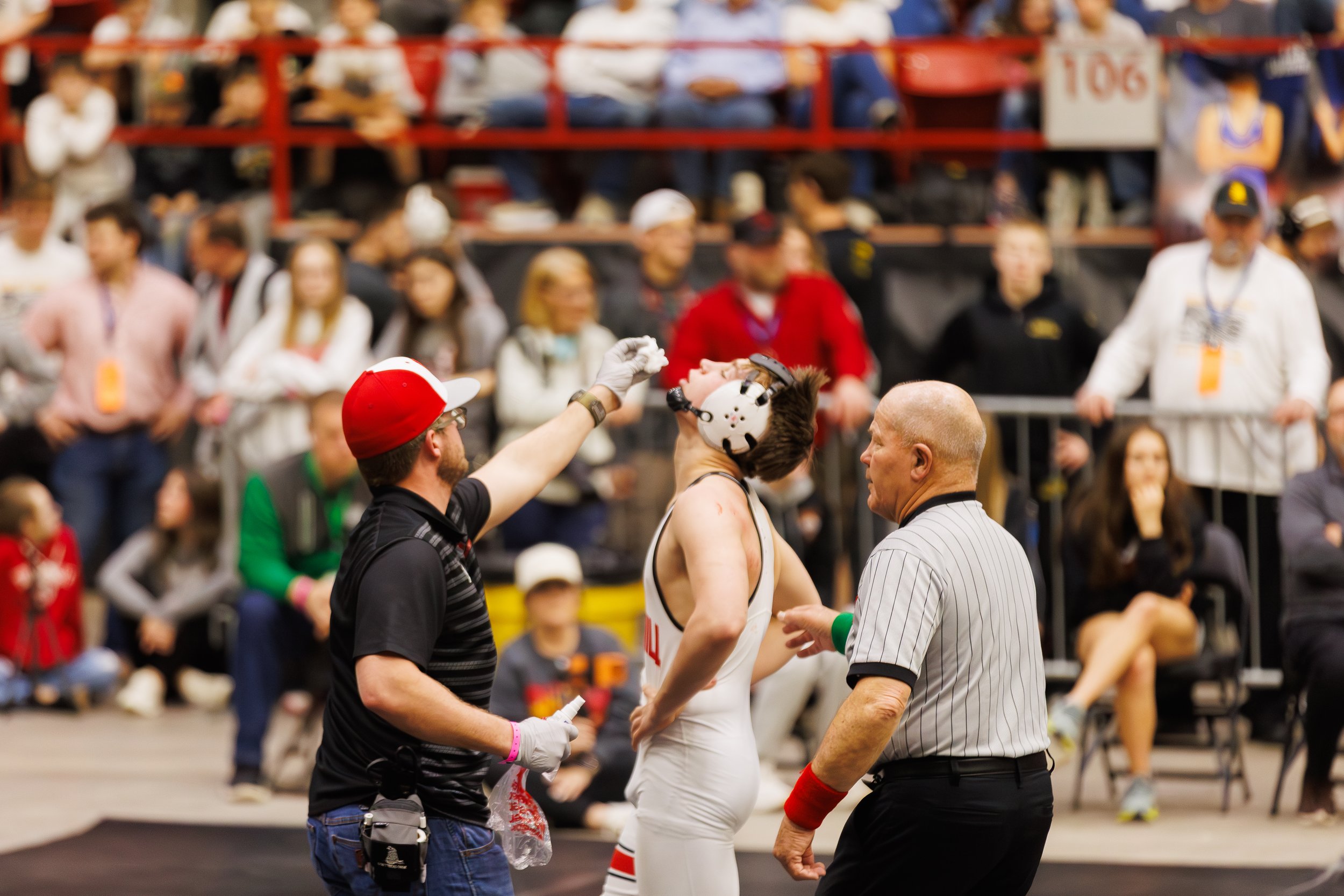 A wrestler in a white uniform and helmet is being attended to by officials after a match stoppage, with spectators in the background.