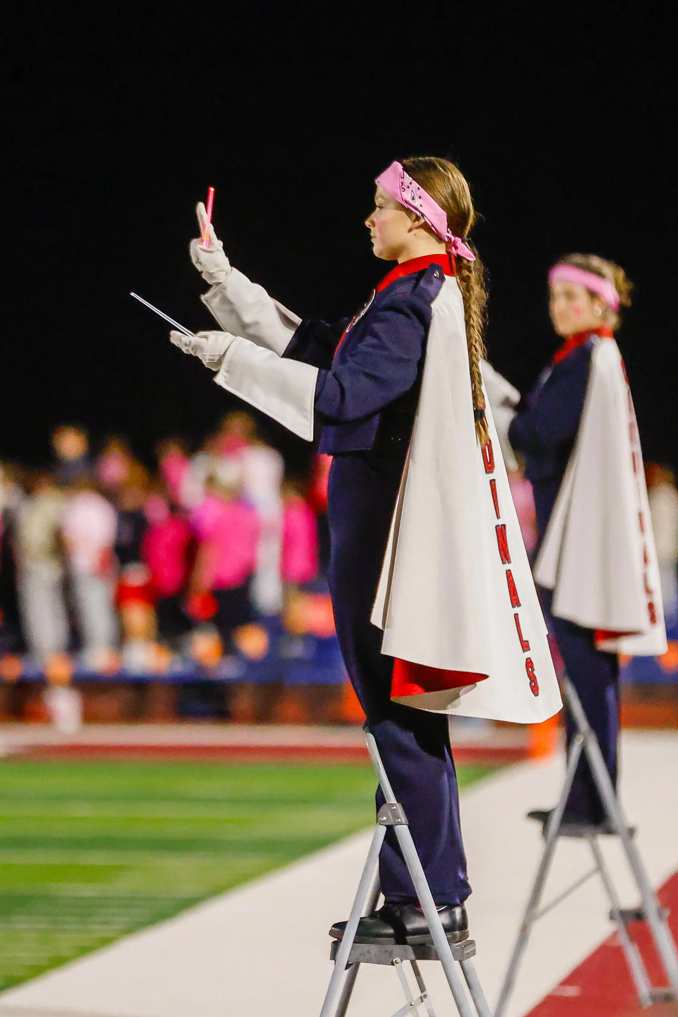 Two female cheerleaders dressed in band uniforms with capes, standing on chairs at a stadium during a night game, with one cheerleader holding a whistle and the other looking at her phone.