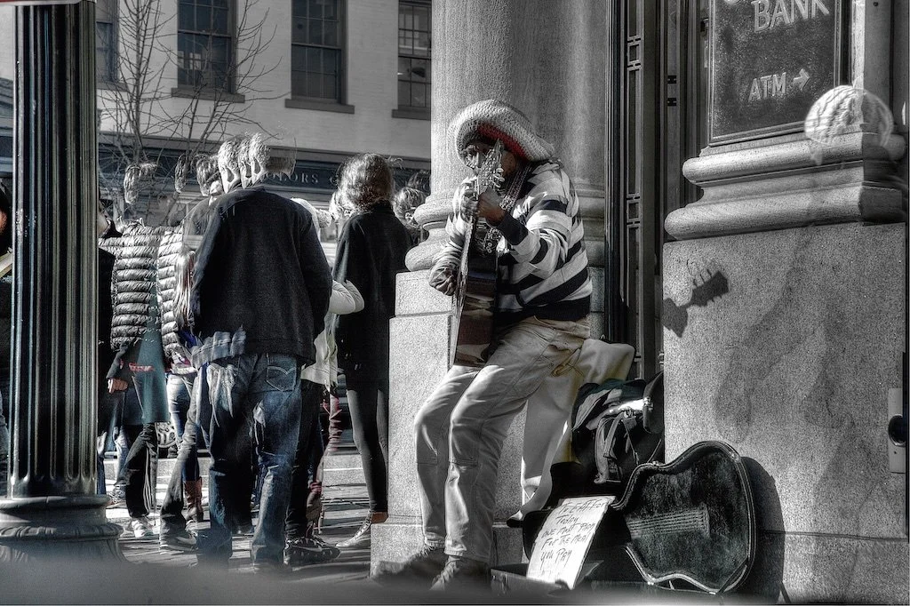 A street performer playing guitar and singing on a city sidewalk, surrounded by pedestrians. The performer is wearing a striped hoodie, white pants, and a hat. A sign and guitar case are on the ground in front of him.