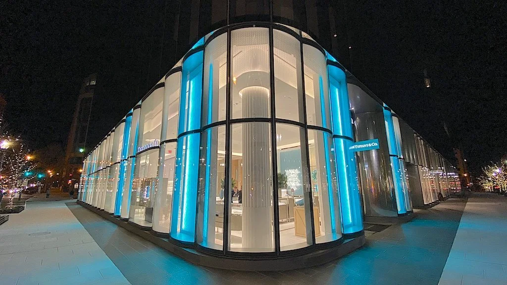 Night view of a modern glass building with the Tiffany & Co. sign, illuminated with blue lights.