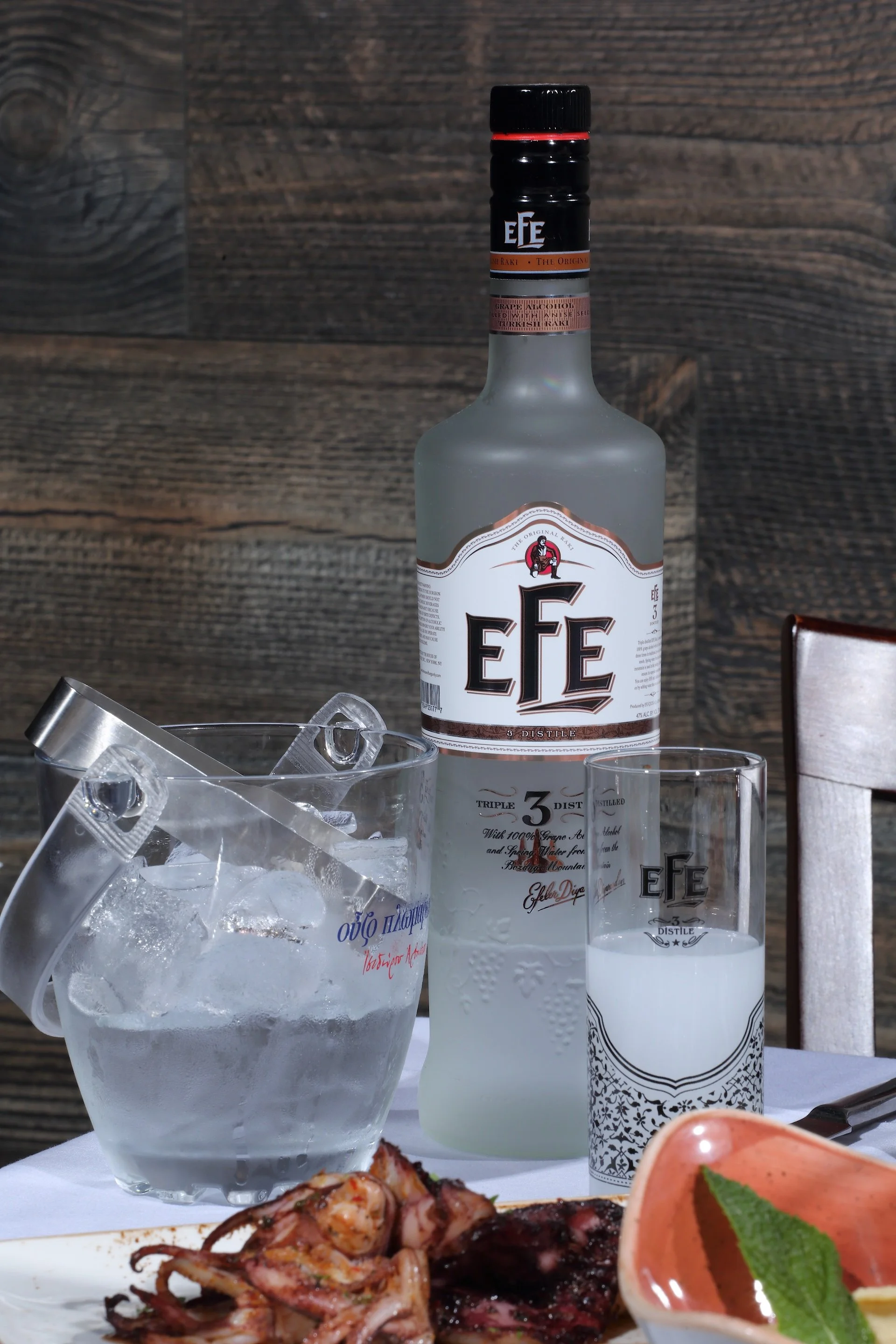 A bottle of EFE Turkish Raki, a glass of milky white raki, a bowl with a green mint leaf, a bowl of grilled octopus, a half slice of lemon, a glass of ice with ice cubes, and a plastic cup with a handle, all placed on a table with a white tablecloth 