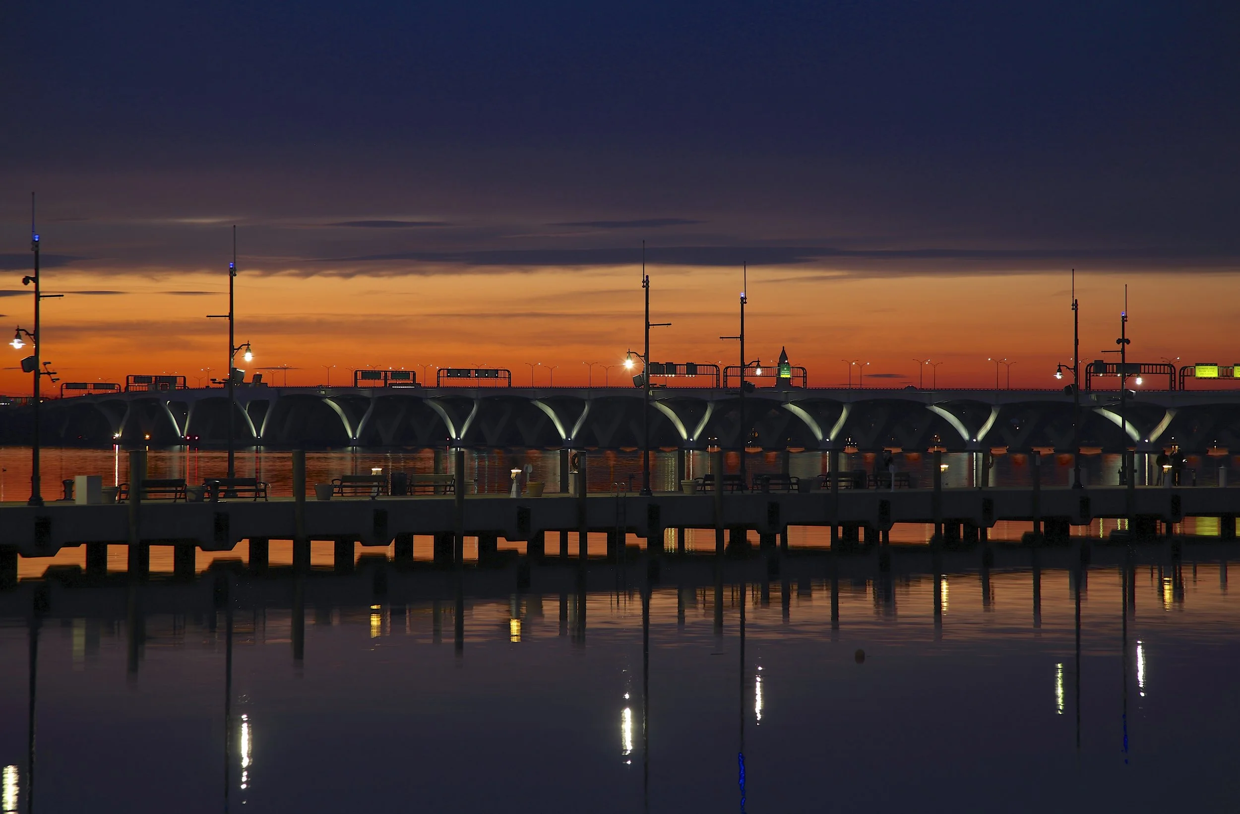 Sunset over a bridge with illuminated street lamps, reflected in calm water below, with a sky transitioning from orange to dark blue.