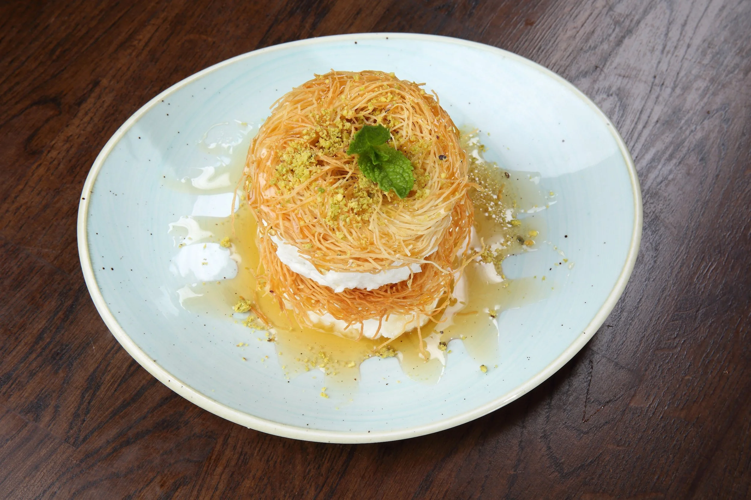 A dessert of two round nests of thin fried noodles, topped with a dollop of white cream or yogurt, garnished with a sprig of mint, and drizzled with syrup, served on a light-colored ceramic plate on a wooden surface.