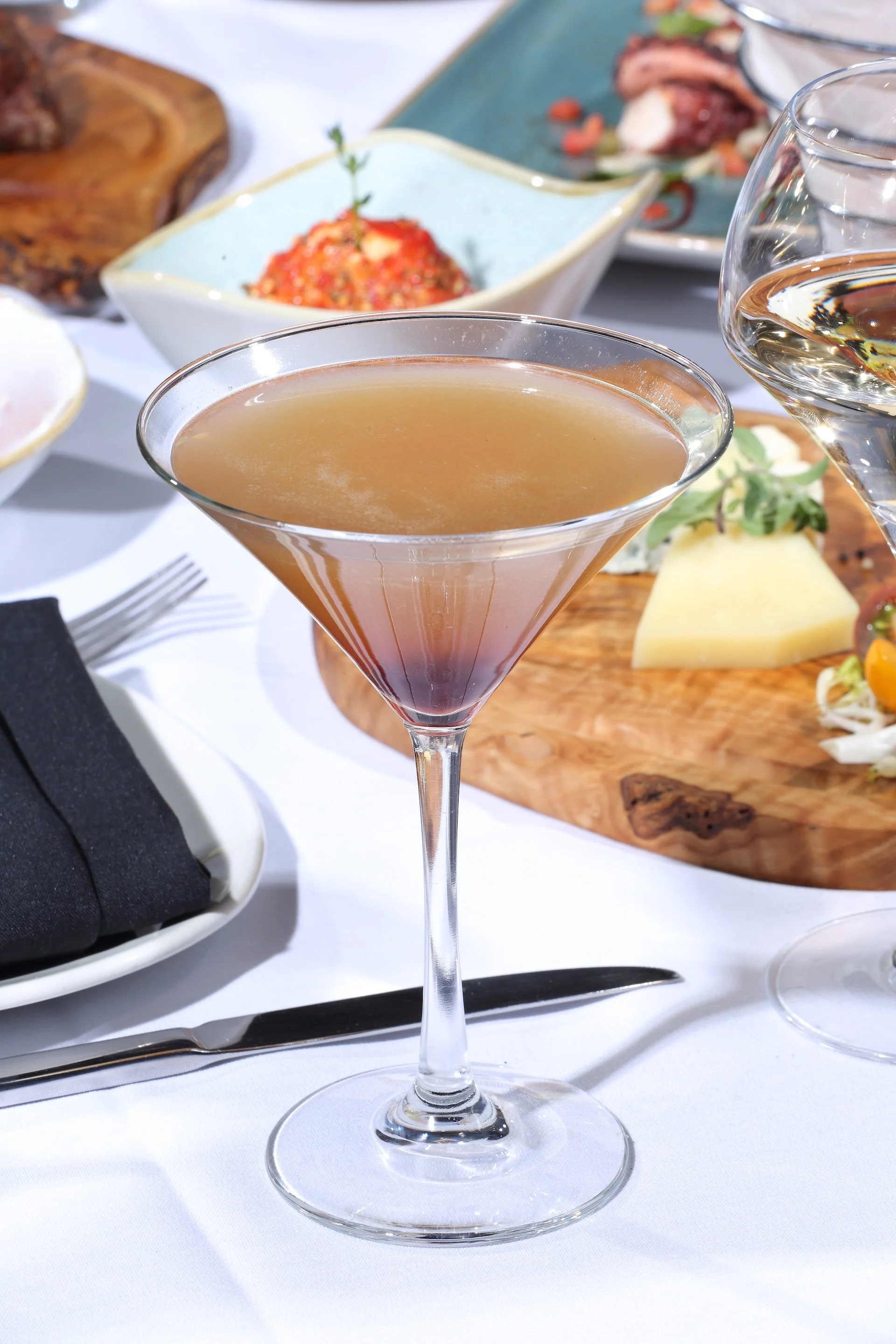 A martini cocktail in a clear stemmed glass on a white tablecloth with other drinks, appetizers, and dishes in the background.
