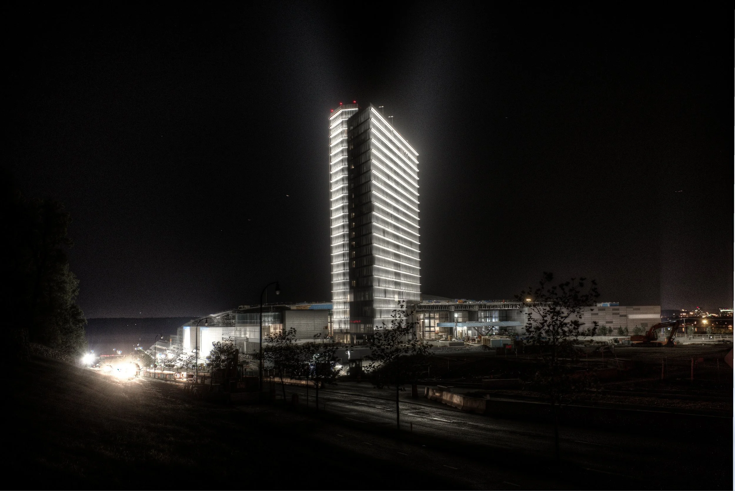 Night view of a tall illuminated MGM Resort Hotel with white lights outlining its edges, situated on a dark cityscape with some illuminated structures and few cars on the road.