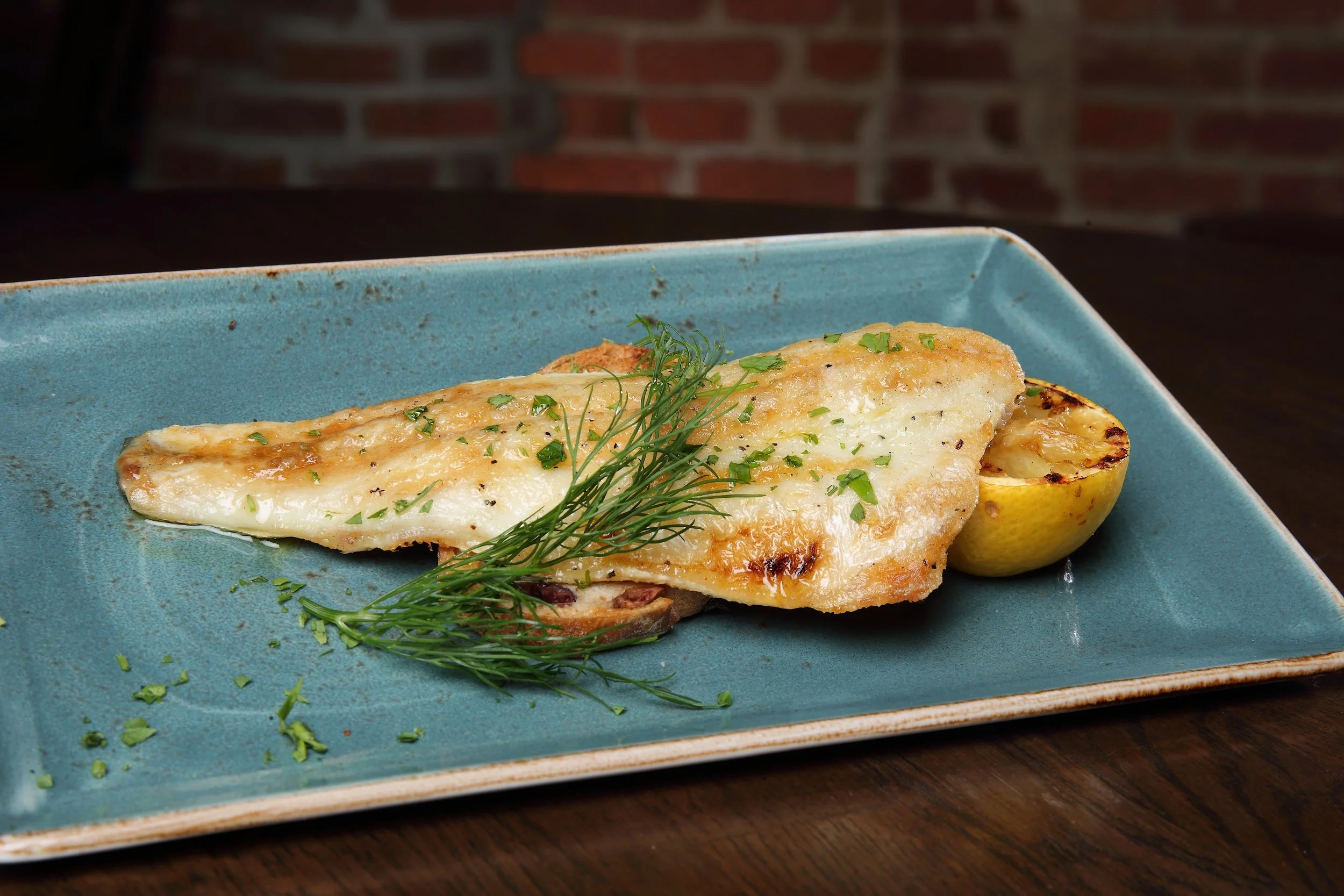 Baked white fish fillet garnished with fresh herbs and served with lemon half on a blue rectangular plate.