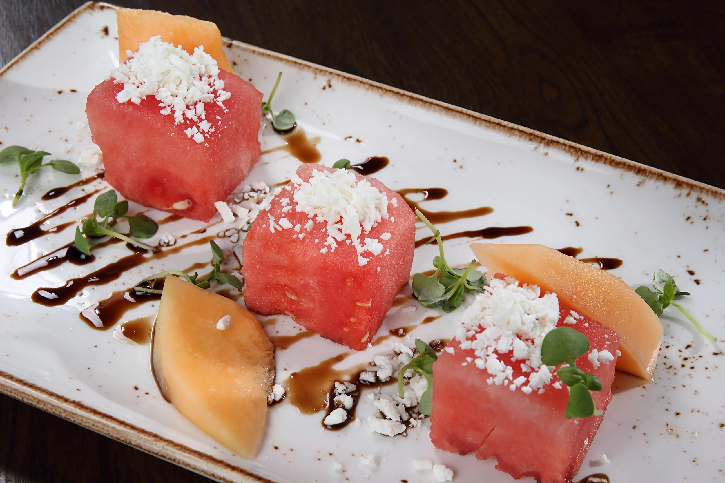 Watermelon and cantaloupe melon cubes topped with white cheese crumbles, garnished with microgreens, drizzled with balsamic reduction, served on a white rectangular plate.