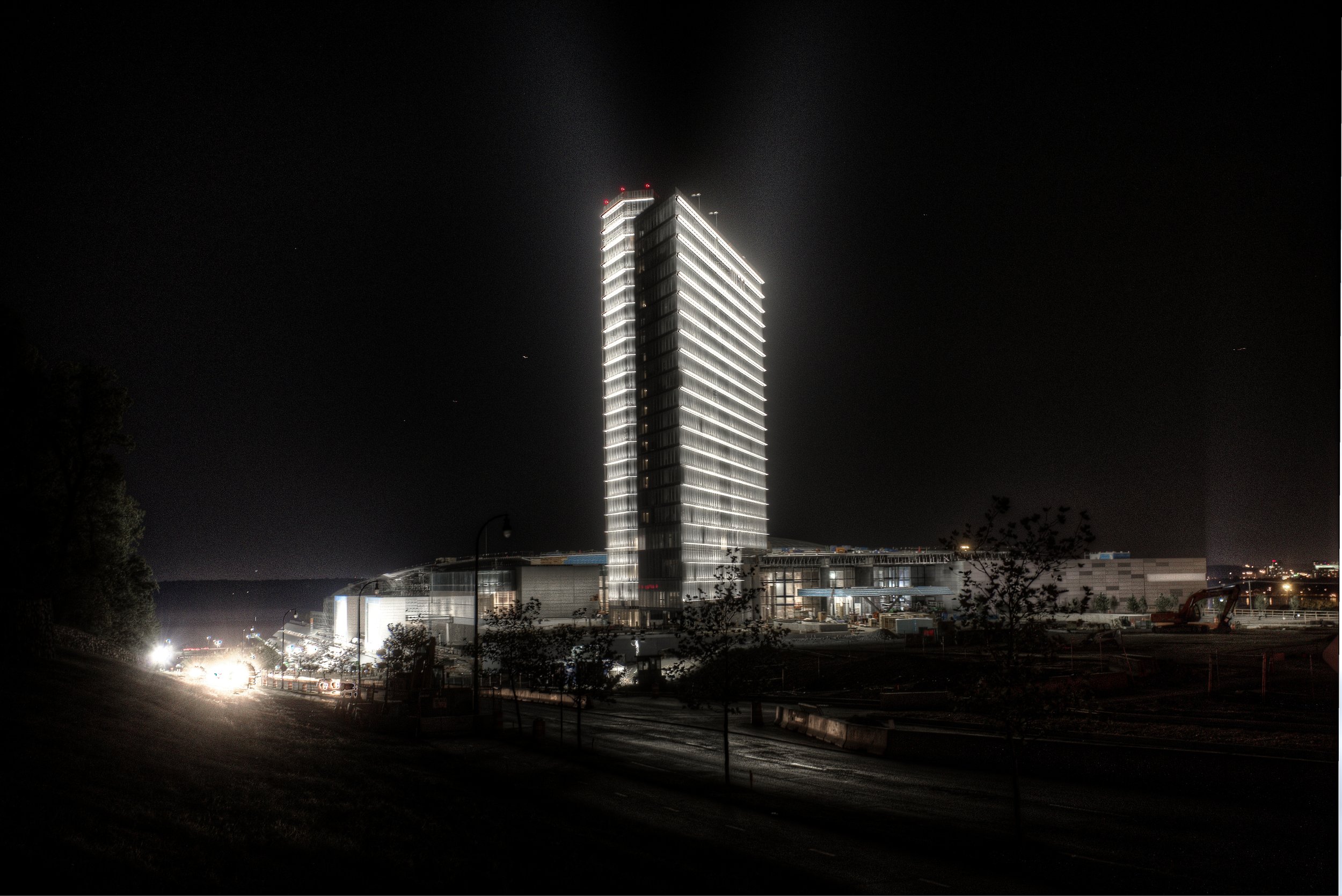 Night view of a tall, illuminated skyscraper with a modern design, next to a large building under construction, with some trees and traffic on the road in the foreground.