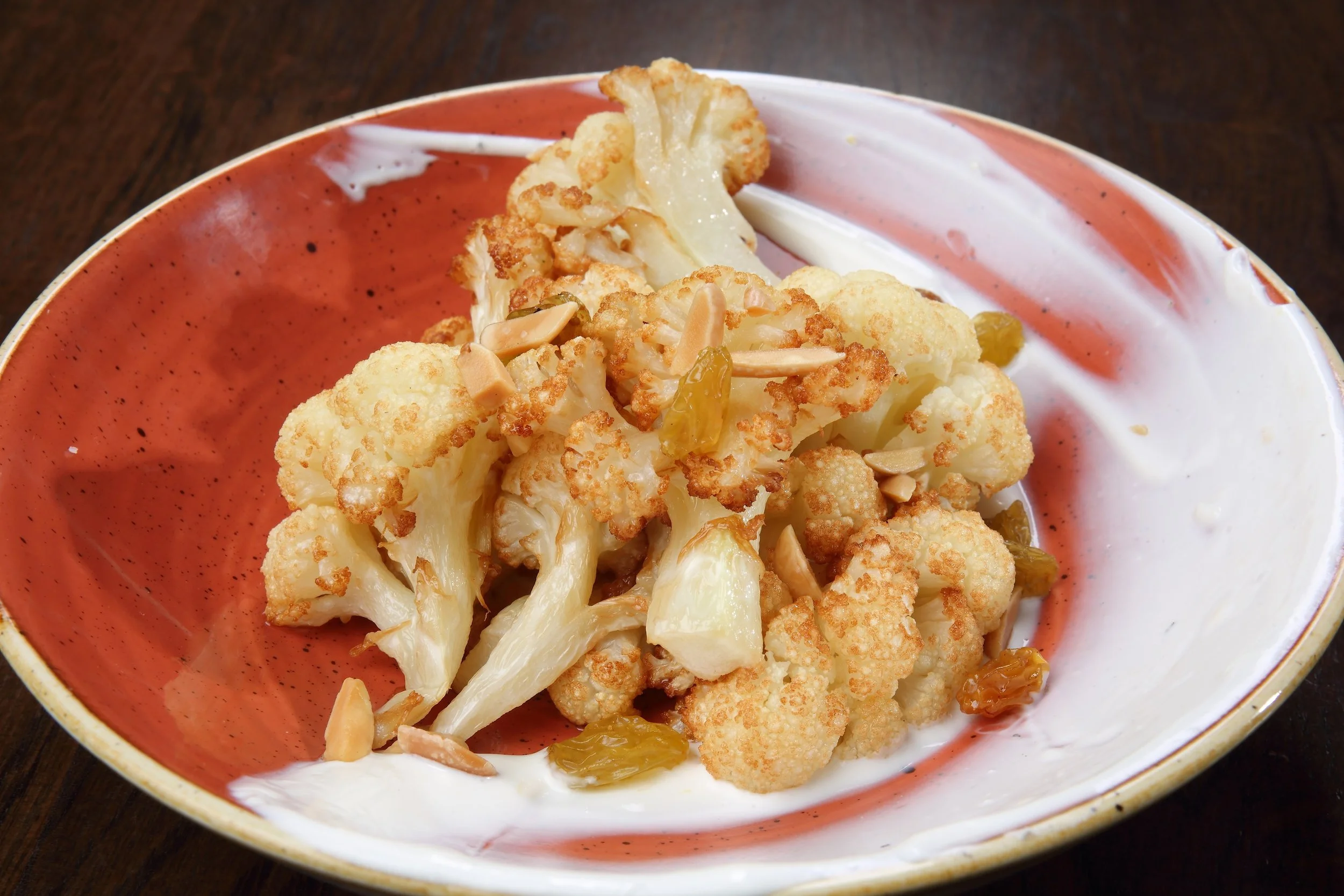 Roasted cauliflower florets topped with sliced almonds and raisins on a colorful ceramic plate with a dollop of Greek yogurt.
