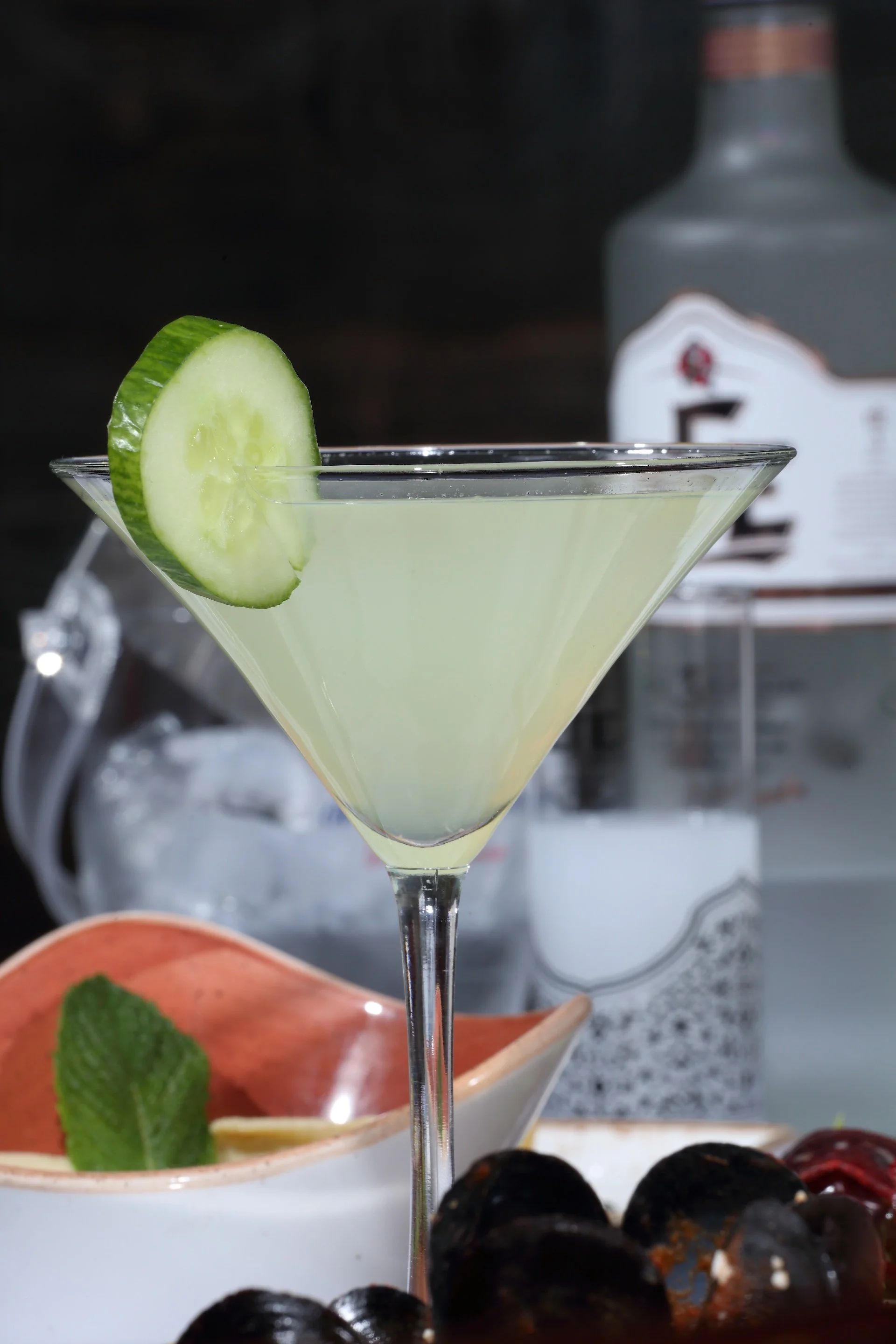 A pale yellow or clear cocktail in a martini glass garnished with a cucumber slice, with a bottle of alcohol, a bowl with lemon and mint, and a cluster of dark shellfish in the background.