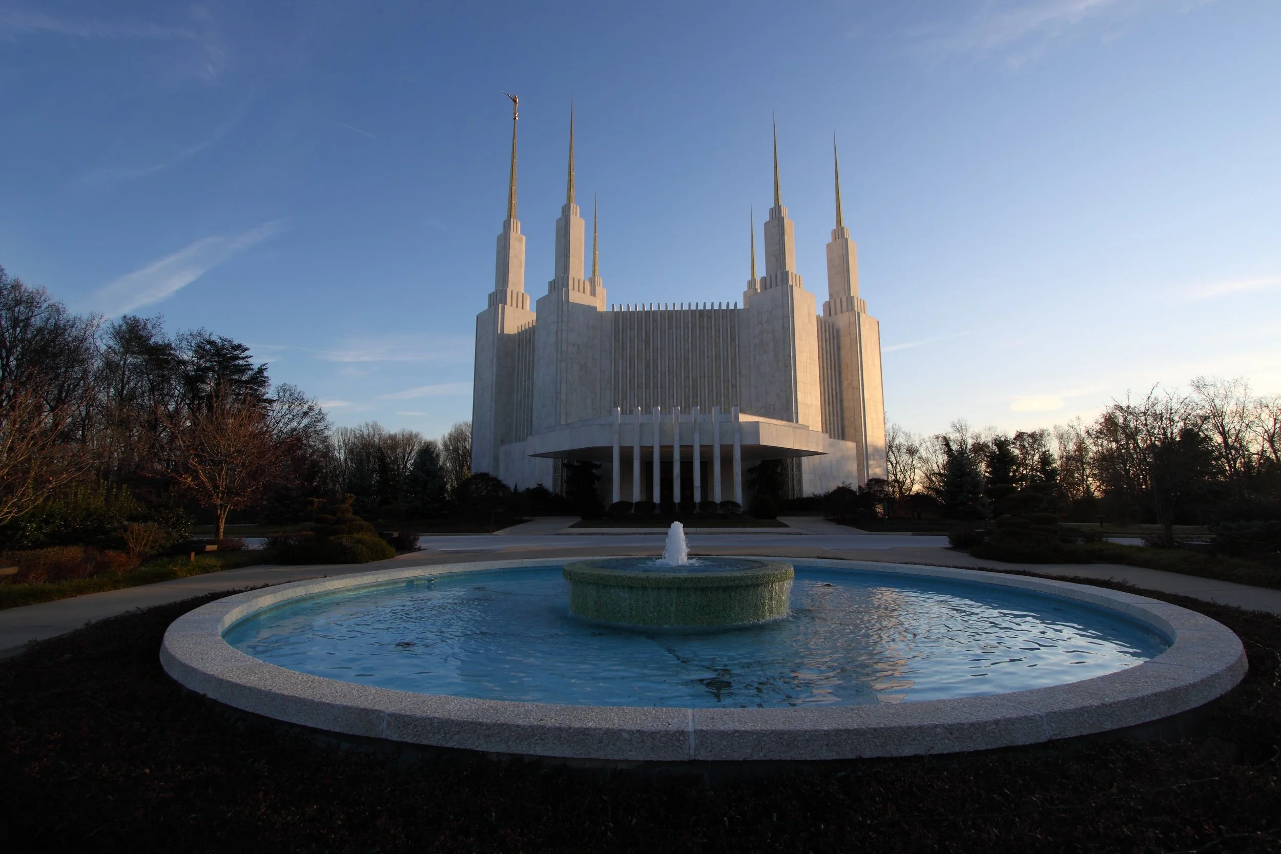 The Salt Lake Temple, a large white Mormon church building with tall spires, is seen in the background with a fountain in the foreground and trees around it during sunset.