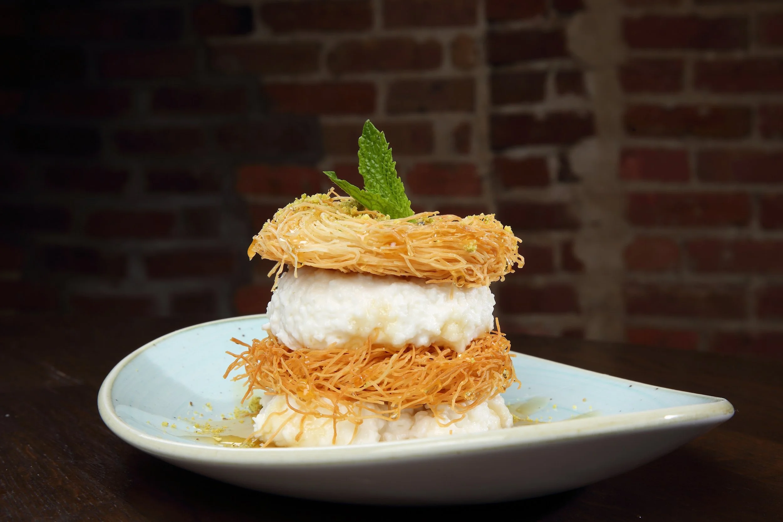 A plated dessert with layers of fried noodles, whipped cream, and a mint leaf garnish on top, set against a dark brick wall background.