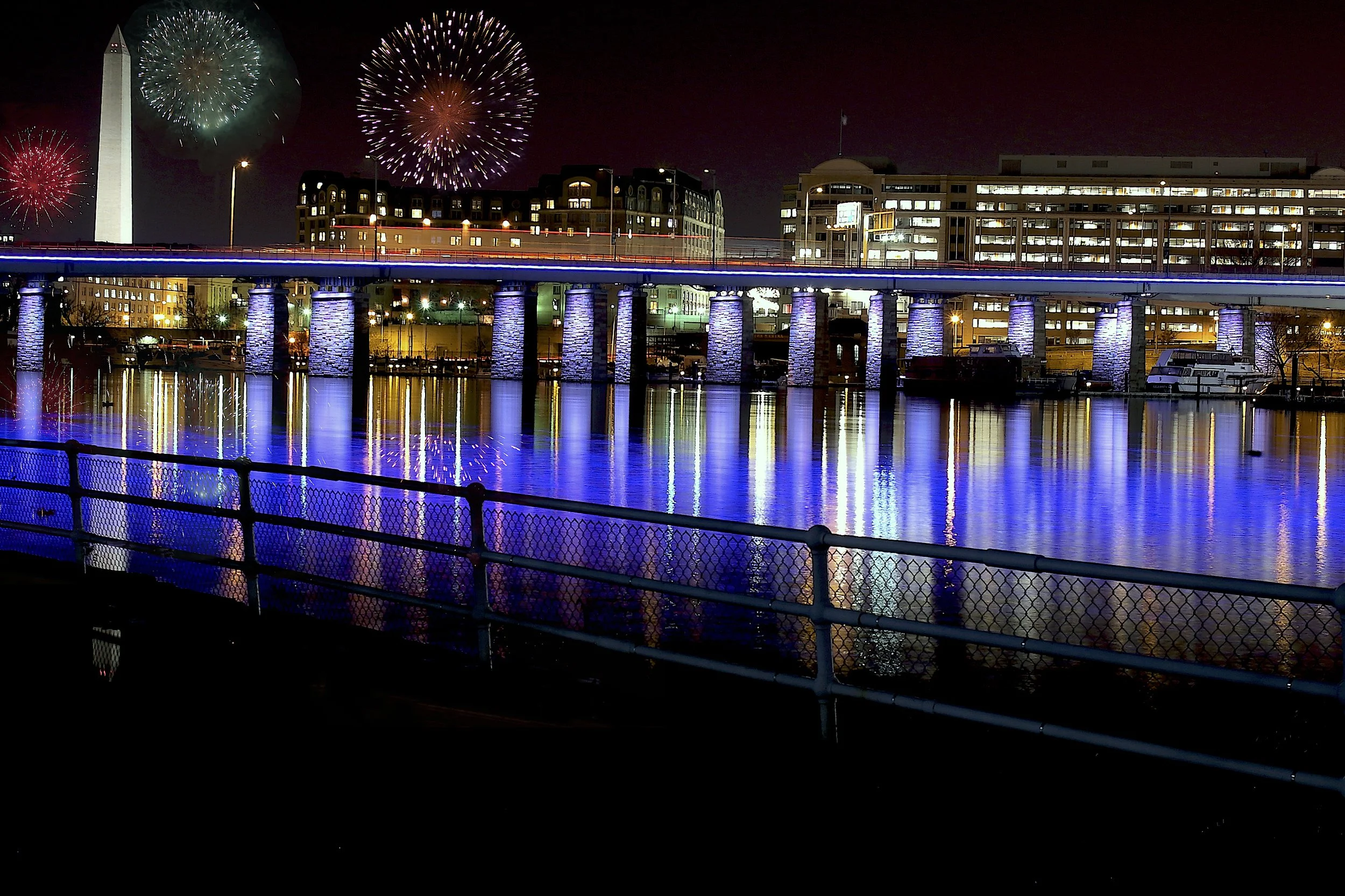 Nighttime scene of the Washington Monument in Washington, D.C.  The Washington Channel reflects the fireworks in the sky, lit bridge reflecting on the water, and illuminated buildings in the background.
