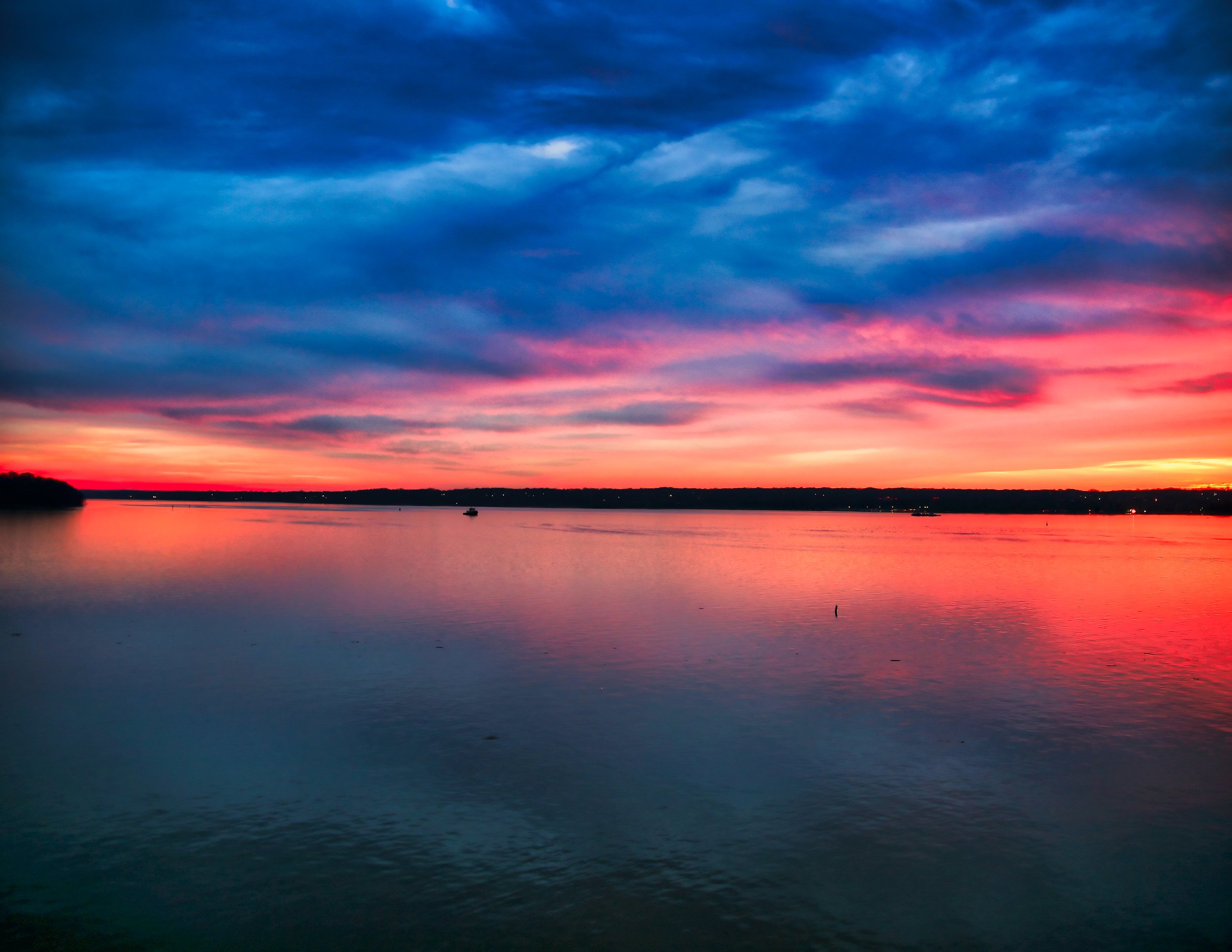 A colorful sunset over a calm body of water with a boat in the distance and a silhouette of land on the horizon.