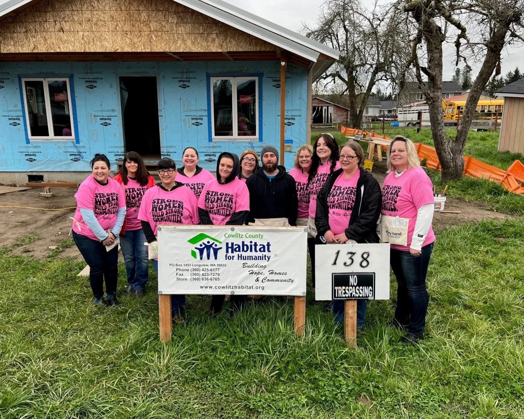 A group of women and one man standing in front of a house under construction, holding a Habitat for Humanity sign. They are wearing pink shirts with the message 'Strong Women Build Strong Communities'. The house behind them is partially built, with some windows installed and the exterior wrapped in blue weatherproof material. There is a tree and some construction materials in the background, along with an orange safety barrier.