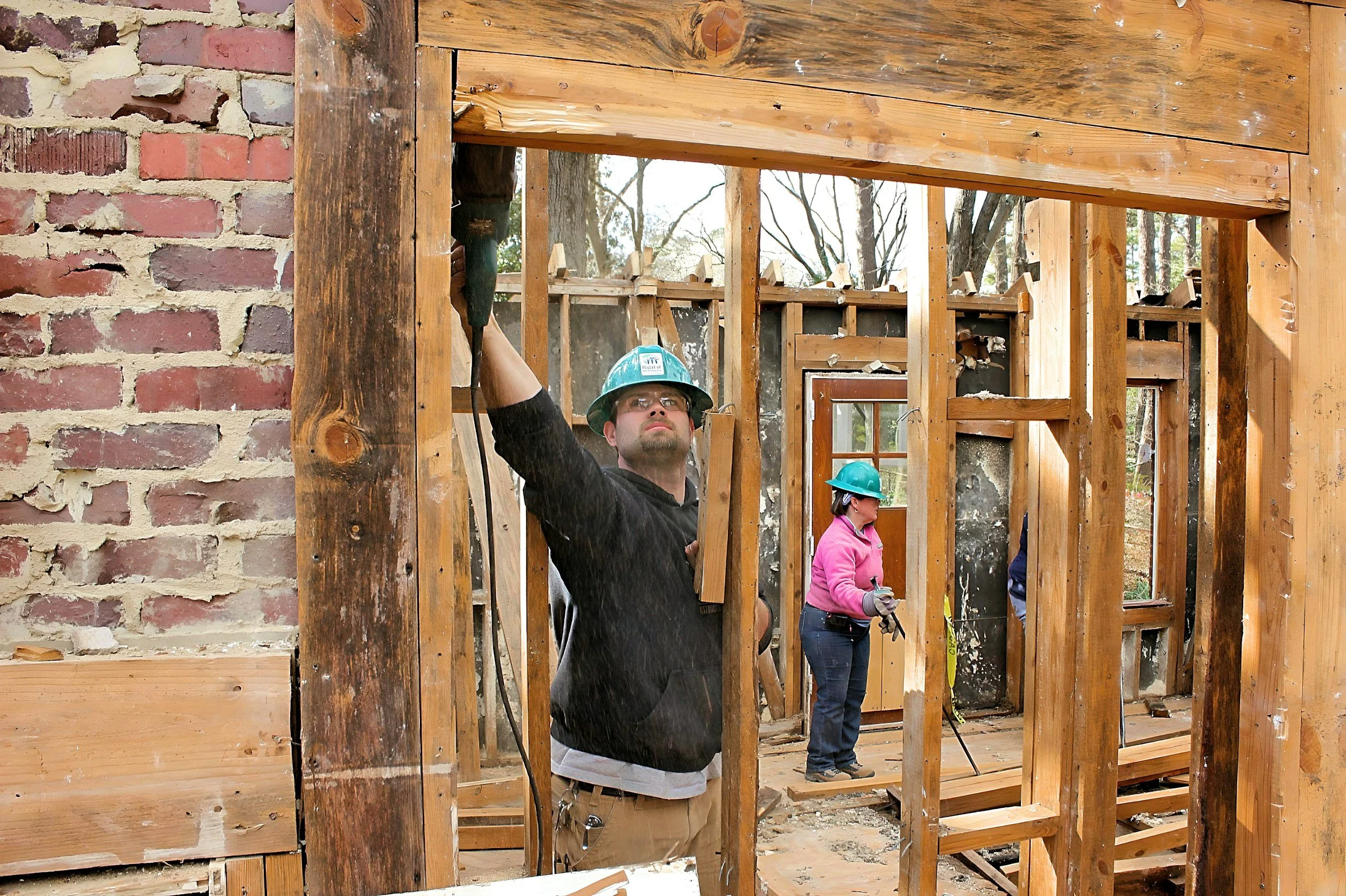 Construction site with wooden framing and brick wall, two workers wearing safety helmets and gloves, one measuring with a tool and the other observing.