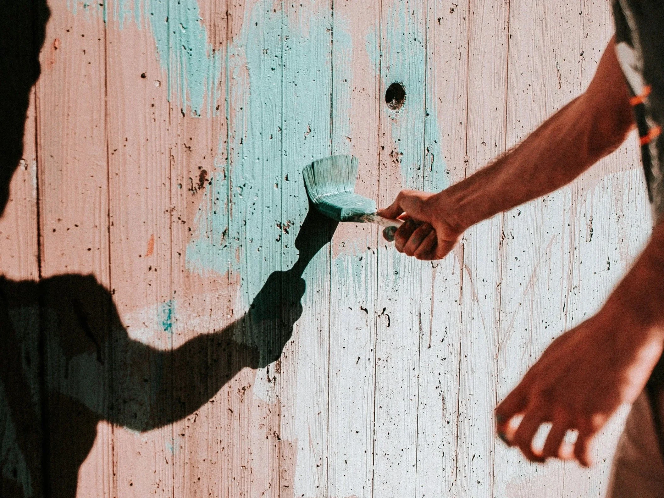 A person painting a wooden surface with light green paint using a paintbrush, with part of another person's hand visible on the right side of the image.
