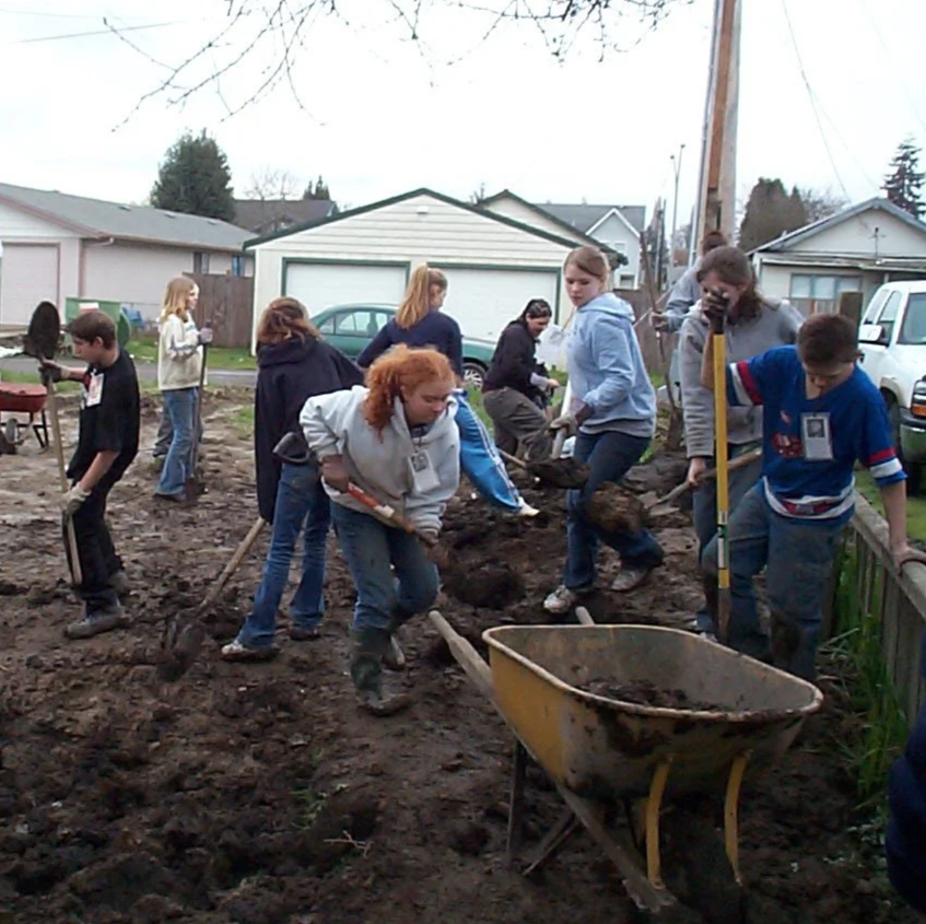 Group of children working together on a garden project, digging in the soil with shovels and wheelbarrow in a residential backyard.