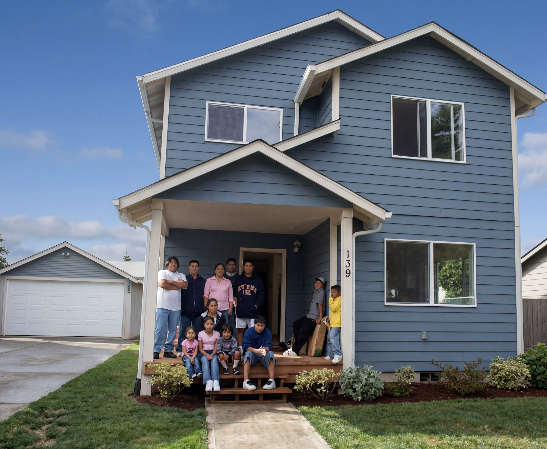 A group of children and adults gathered on the front porch of a blue two-story house with a white garage in the background. The house has a small garden with shrubs and a walkway leading to the porch.