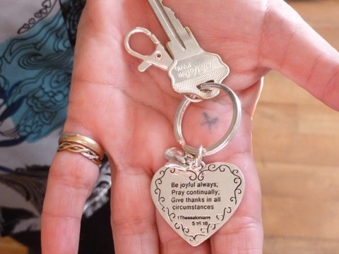 Hand holding a set of keys with a heart-shaped keychain engraved with a prayer and a small star tattoo on the palm.