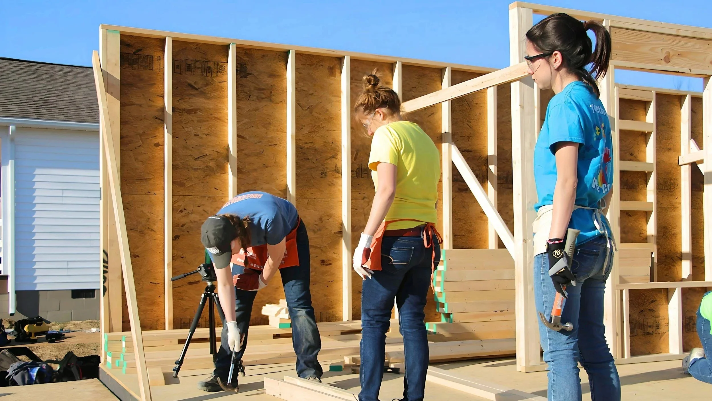 People constructing a wooden house, with tools and equipment on-site, in a clear outdoor setting.