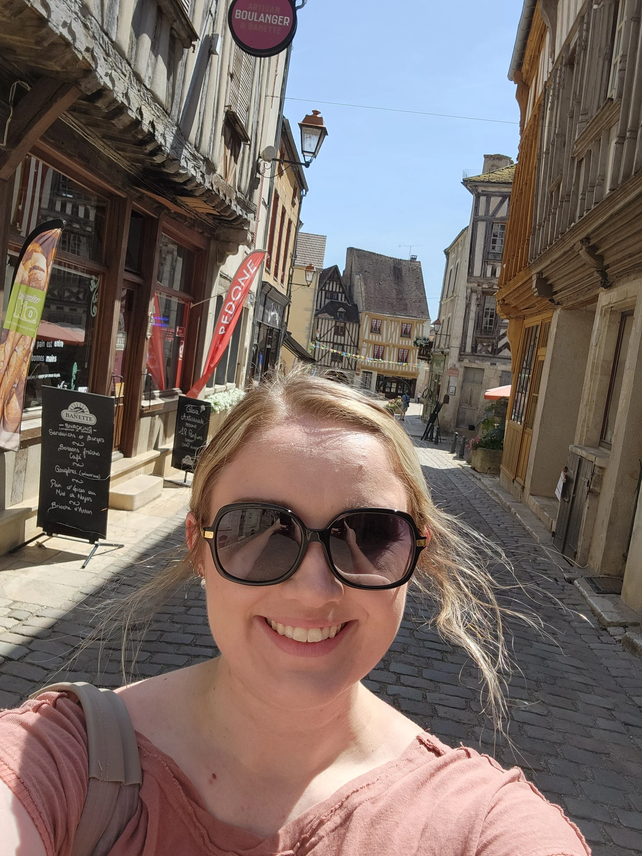 A woman with blonde hair wearing large sunglasses and a pink top taking a selfie on a cobblestone street in a European town with historic buildings and storefronts.