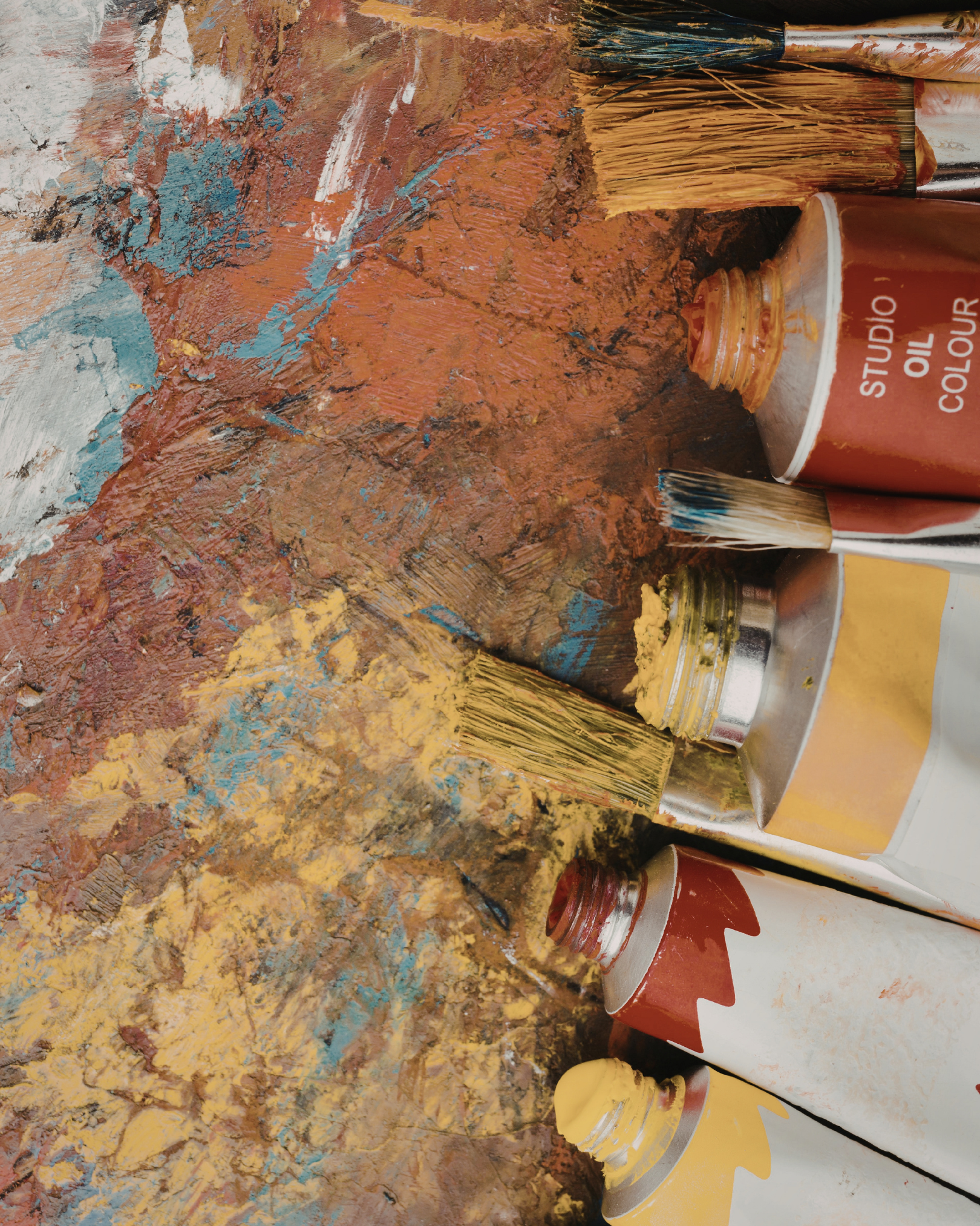Artists' paint brushes, tubes of yellow and brown acrylic paint, and a can of studio oil color on a textured, multicolored abstract painted surface.