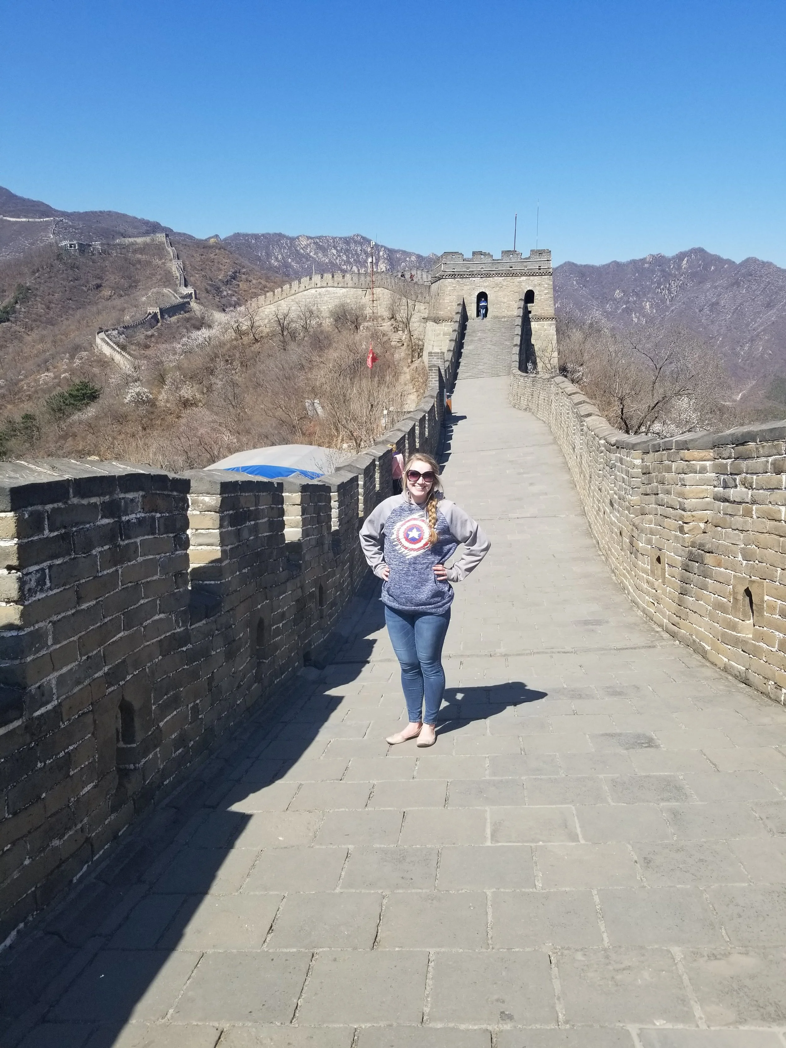A woman wearing sunglasses, a Captain America hoodie, and jeans posing with hands on hips on the Great Wall of China under a clear blue sky.