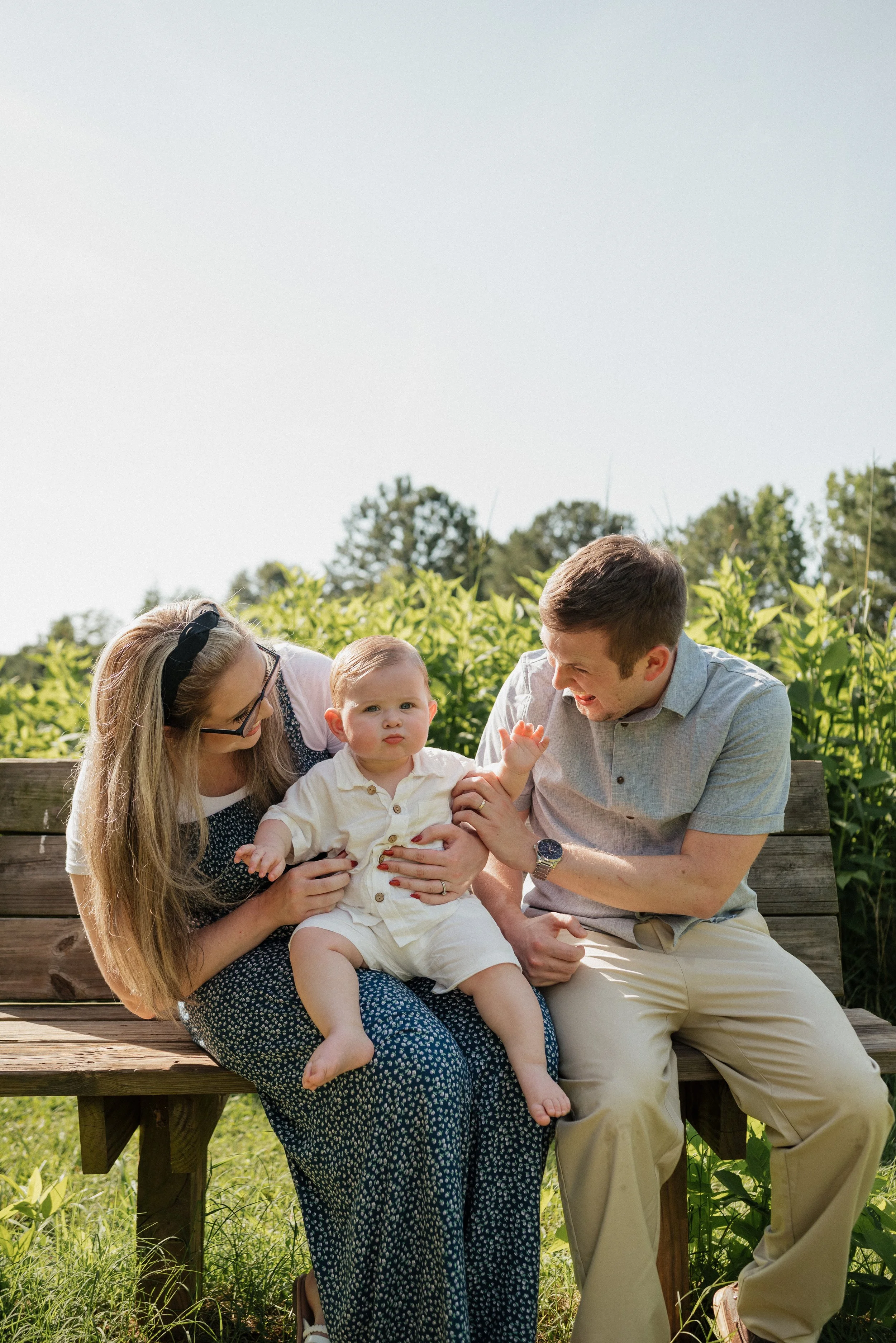 Family of three sitting on a wooden bench outdoors, with greenery and trees behind them. The mother and father are playing with a young boy on their laps. The family appears to be enjoying a sunny day.