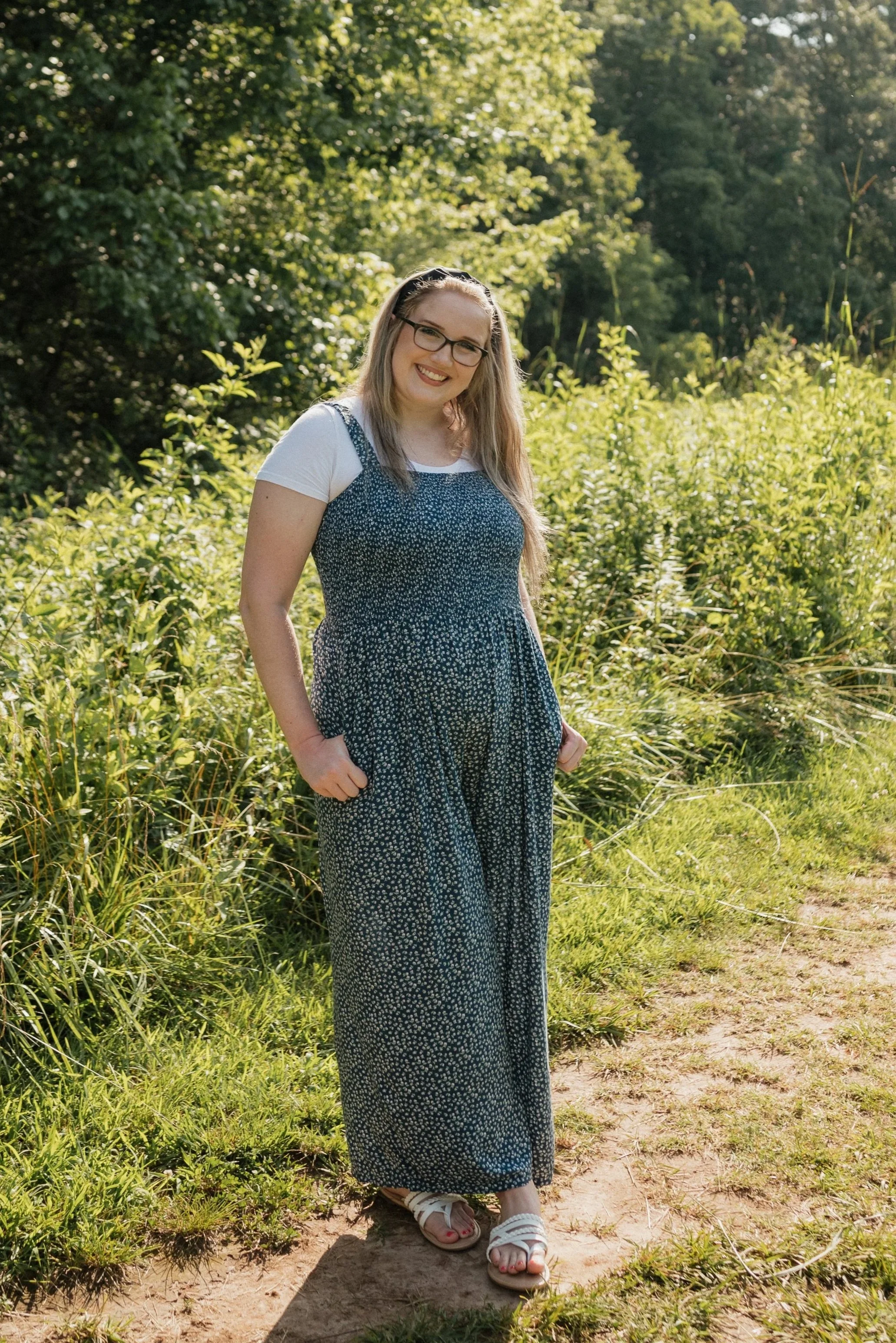 A woman with glasses and long blonde hair, wearing a white t-shirt under a blue floral jumpsuit and white sandals, standing outdoors on a dirt path surrounded by green shrubs and trees.