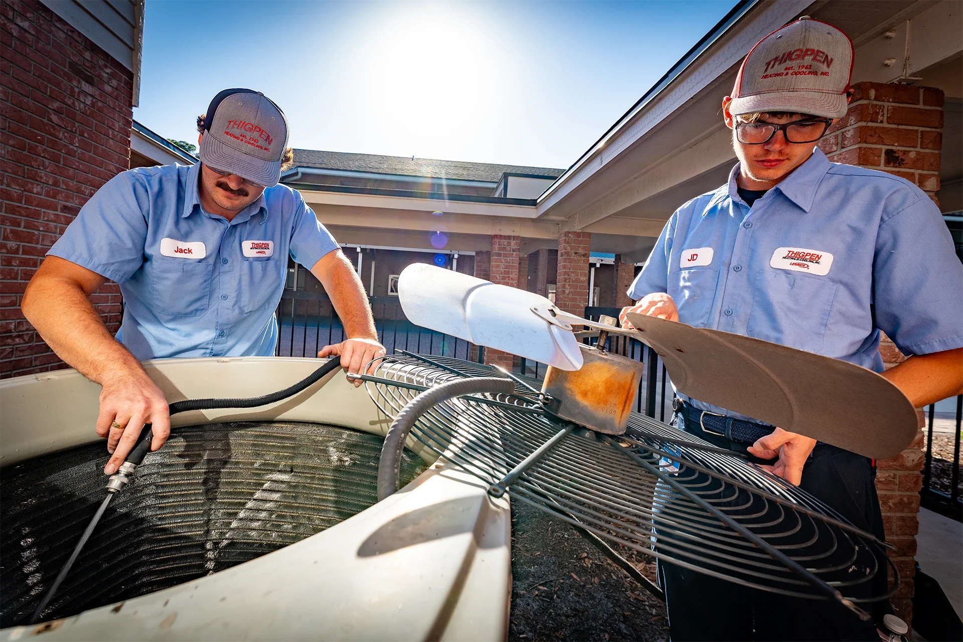 Two HVAC technicians wearing gray hats and blue shirts working on an air conditioning unit outside a brick building.