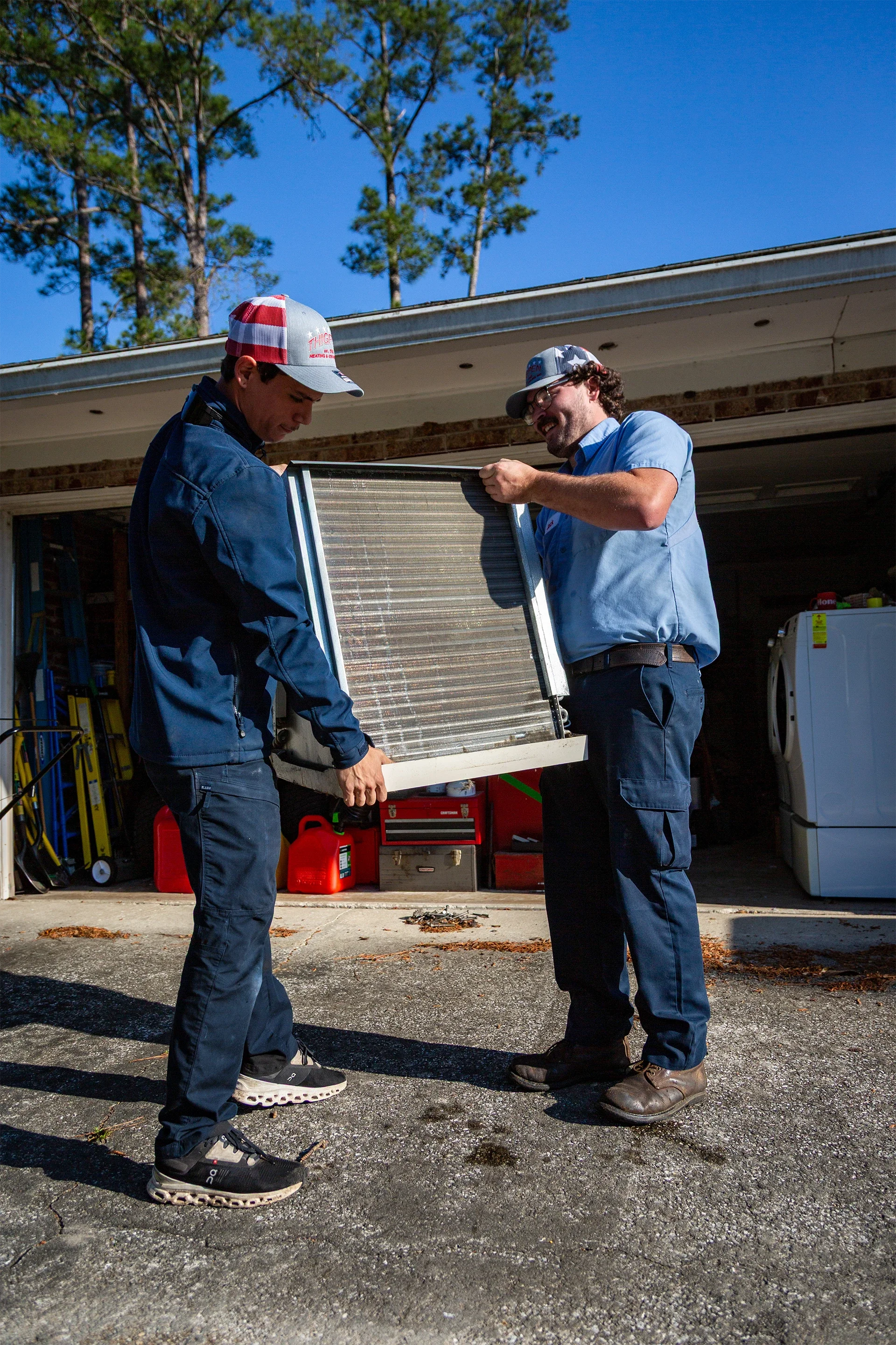 Two men in work uniforms inspecting a dirty air filter in a driveway, with garage tools and storage in the background.