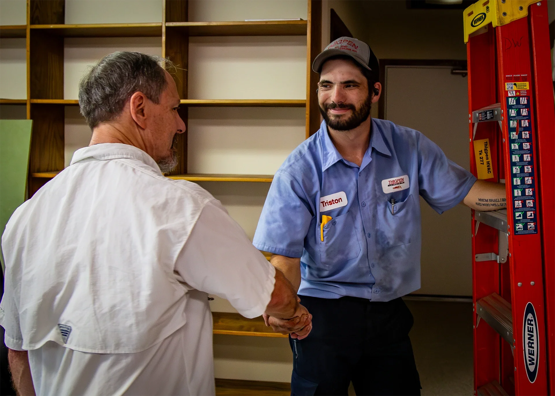 A man with a beard wearing a blue shirt and gray cap shaking hands with an elderly man wearing a white shirt, in a room with wooden shelves and a yellow and red ladder.