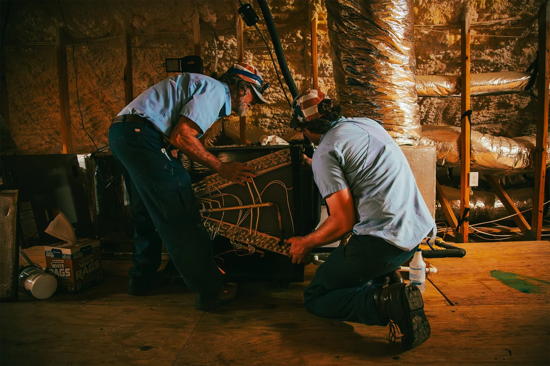 Two men working on a large machine inside a wooden room with insulation on the walls, with boxes of trash bags and a bottle nearby.