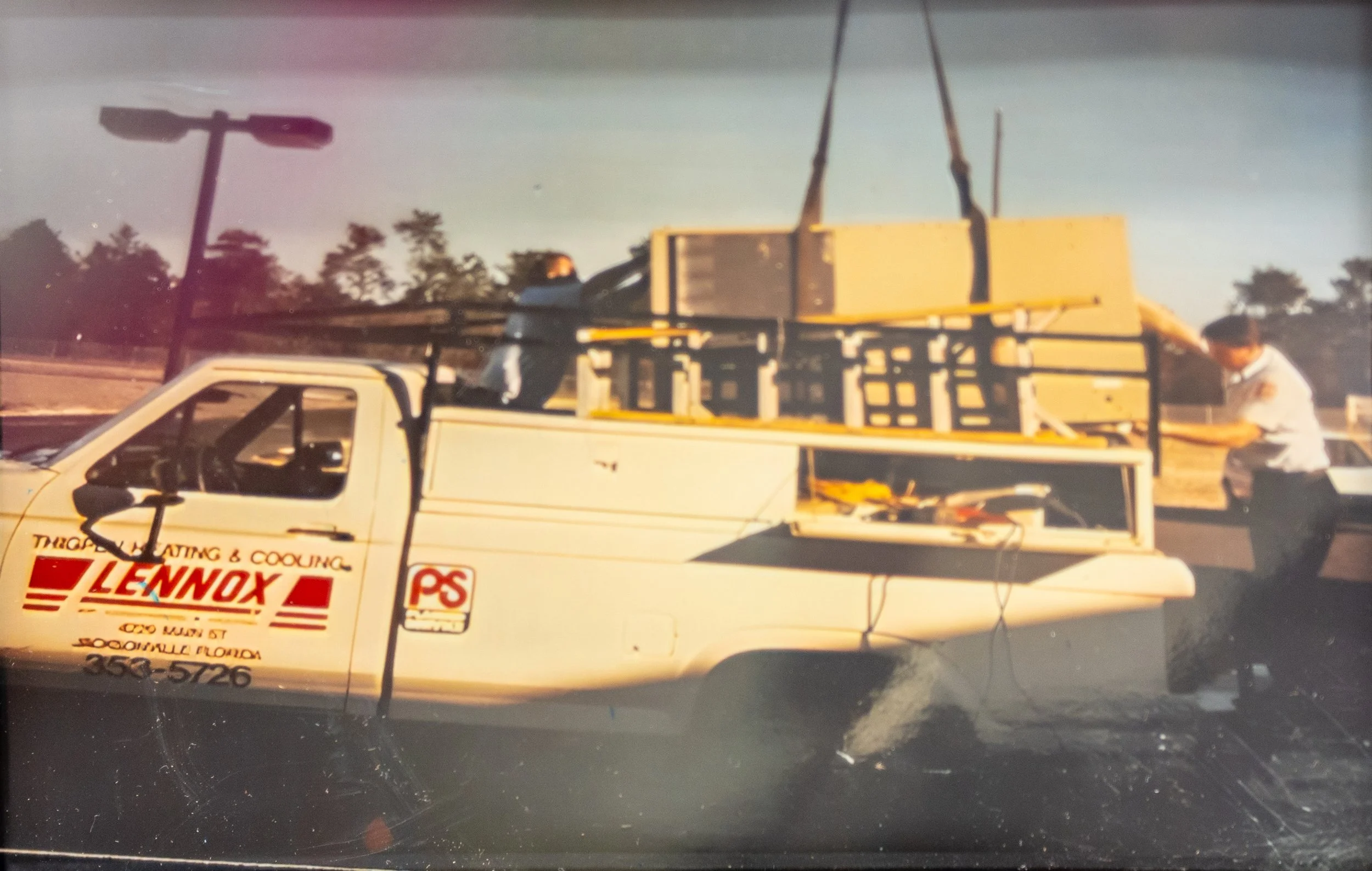 A white pickup truck with advertisements on the side, a mounted yellow work platform, and workers on the truck bed working on the platform in an outdoor parking lot.
