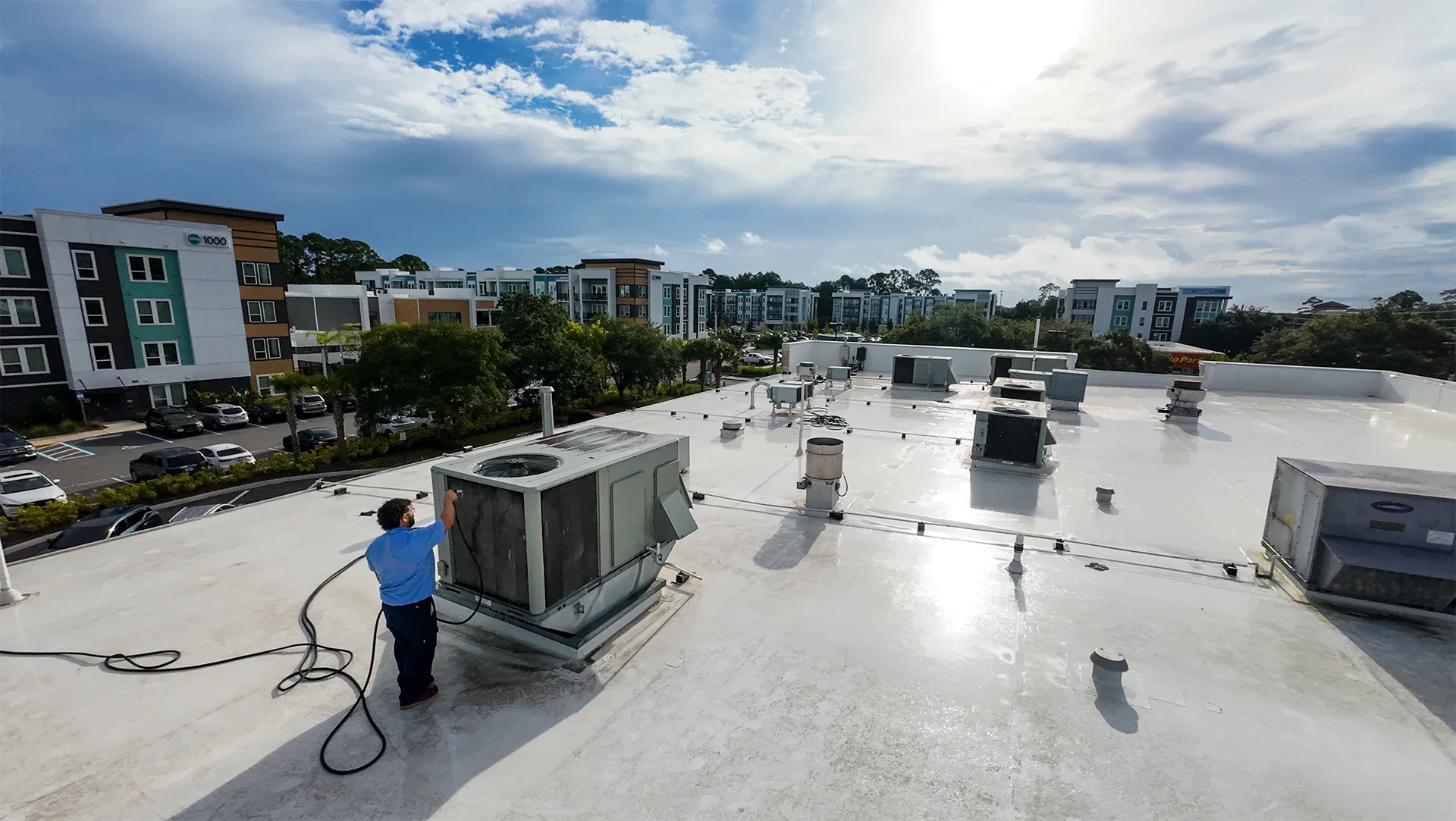 A man in a blue shirt working on HVAC units on a flat rooftop in a residential or commercial area with apartment buildings and parking lot in the background under partly cloudy sky.
