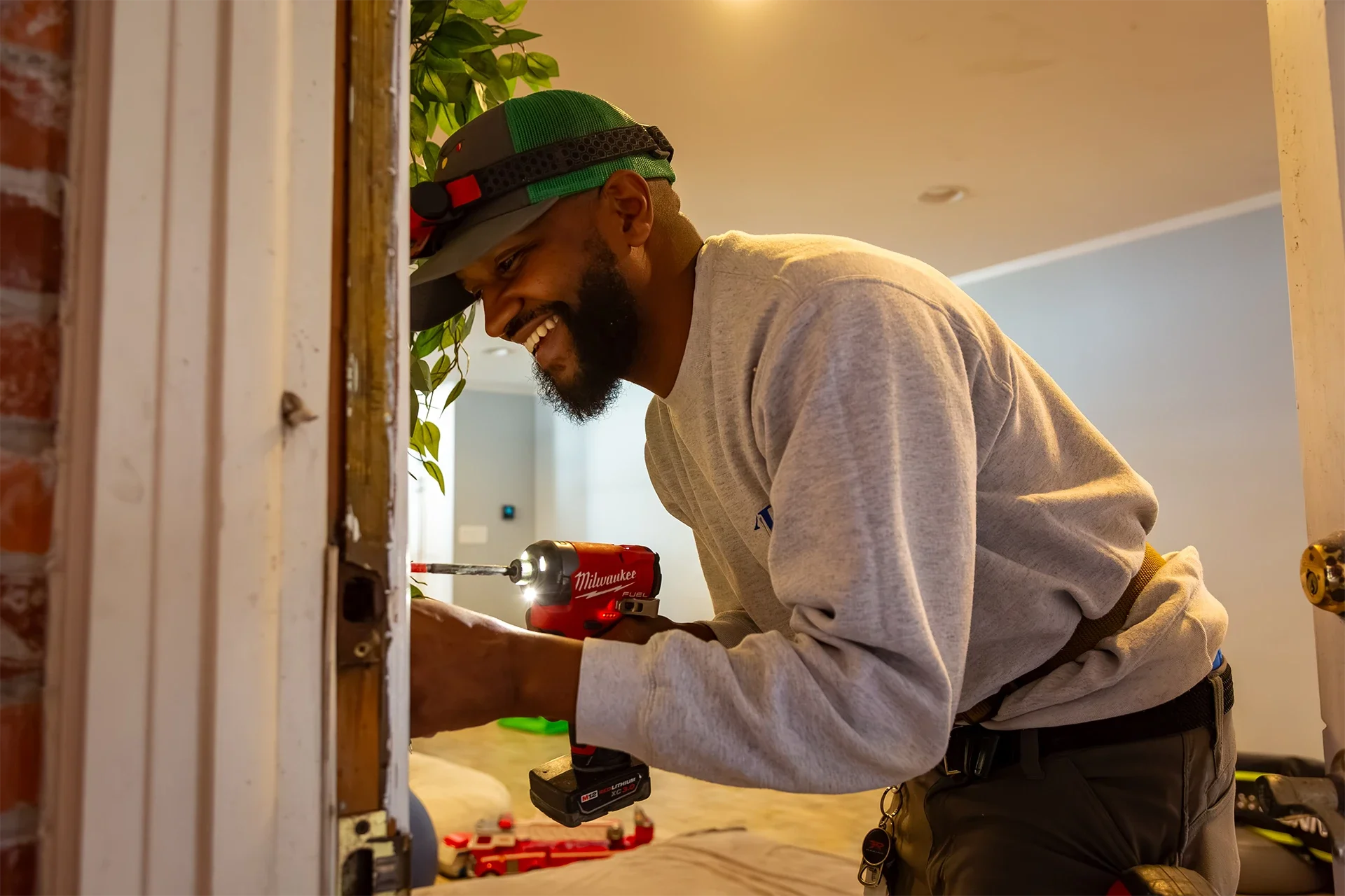 A man wearing a baseball cap, grey shirt, and black pants is working on a home improvement project using a cordless drill with a red body. He is smiling and focused on the task.