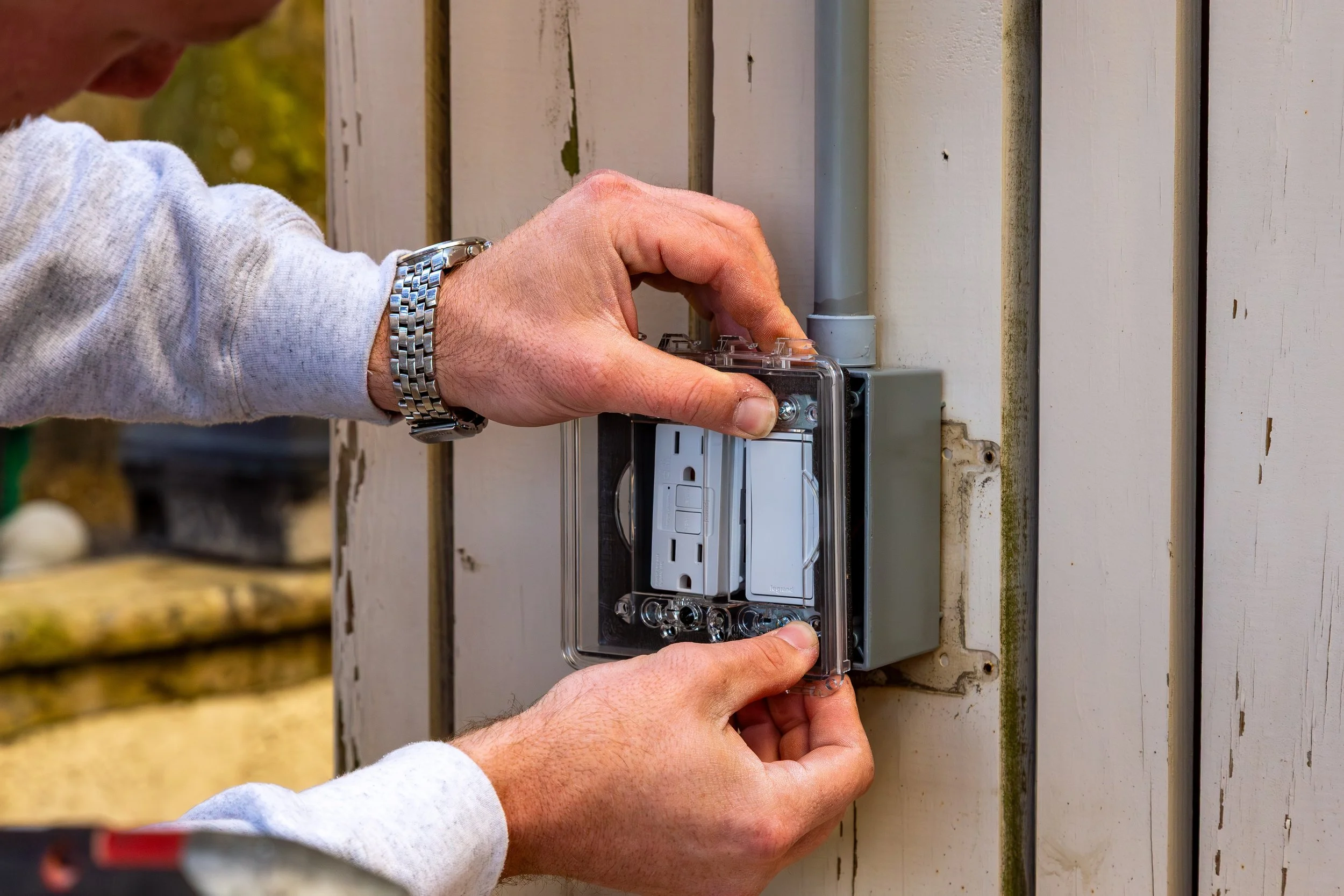 Person installing or fixing an electrical outlet on an outdoor wall, wearing a gray sweatshirt and a silver wristwatch.