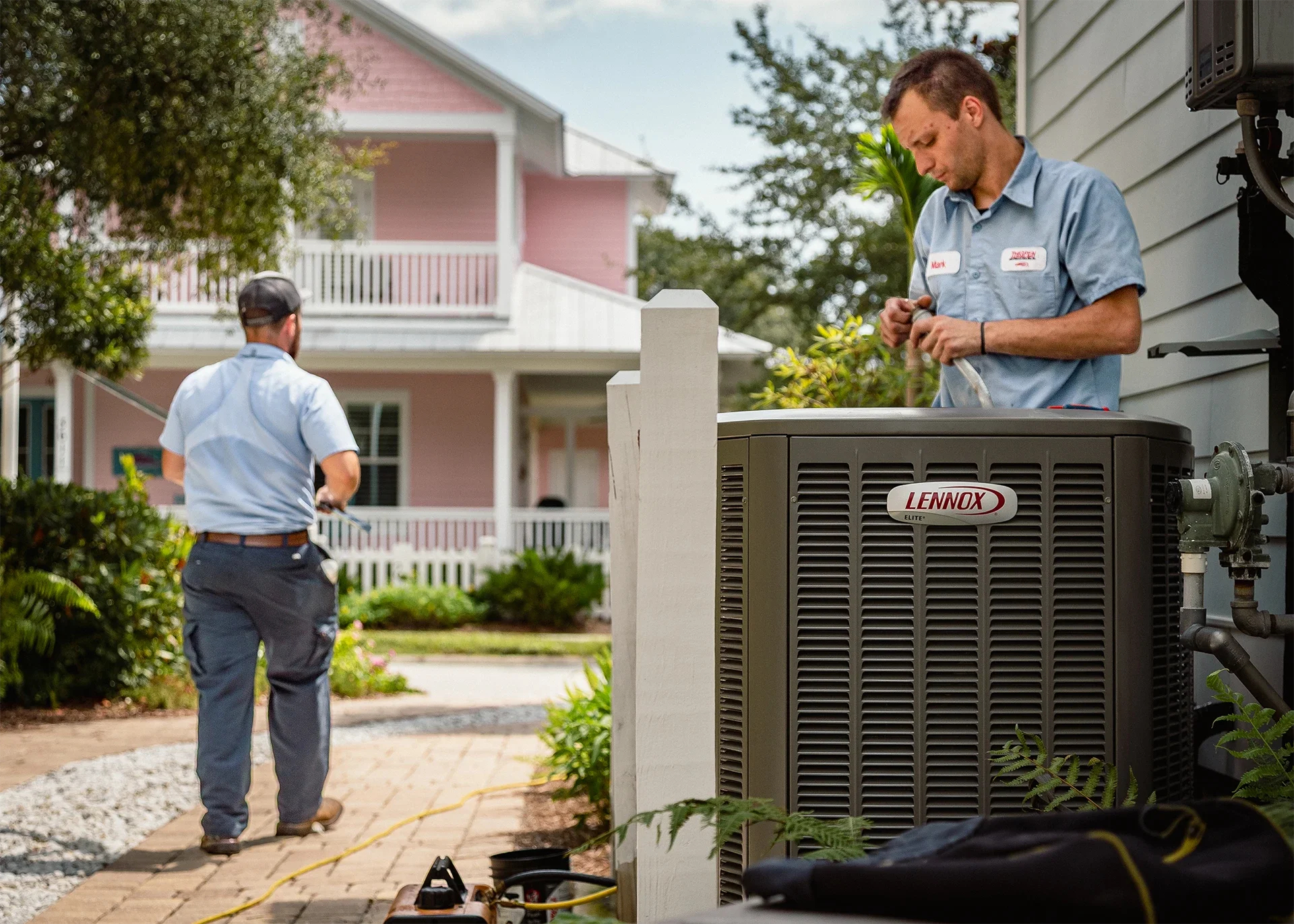 Two HVAC technicians working on an air conditioning unit outside a house. One is on the ground, and the other is on the right side, adjusting something on the unit.